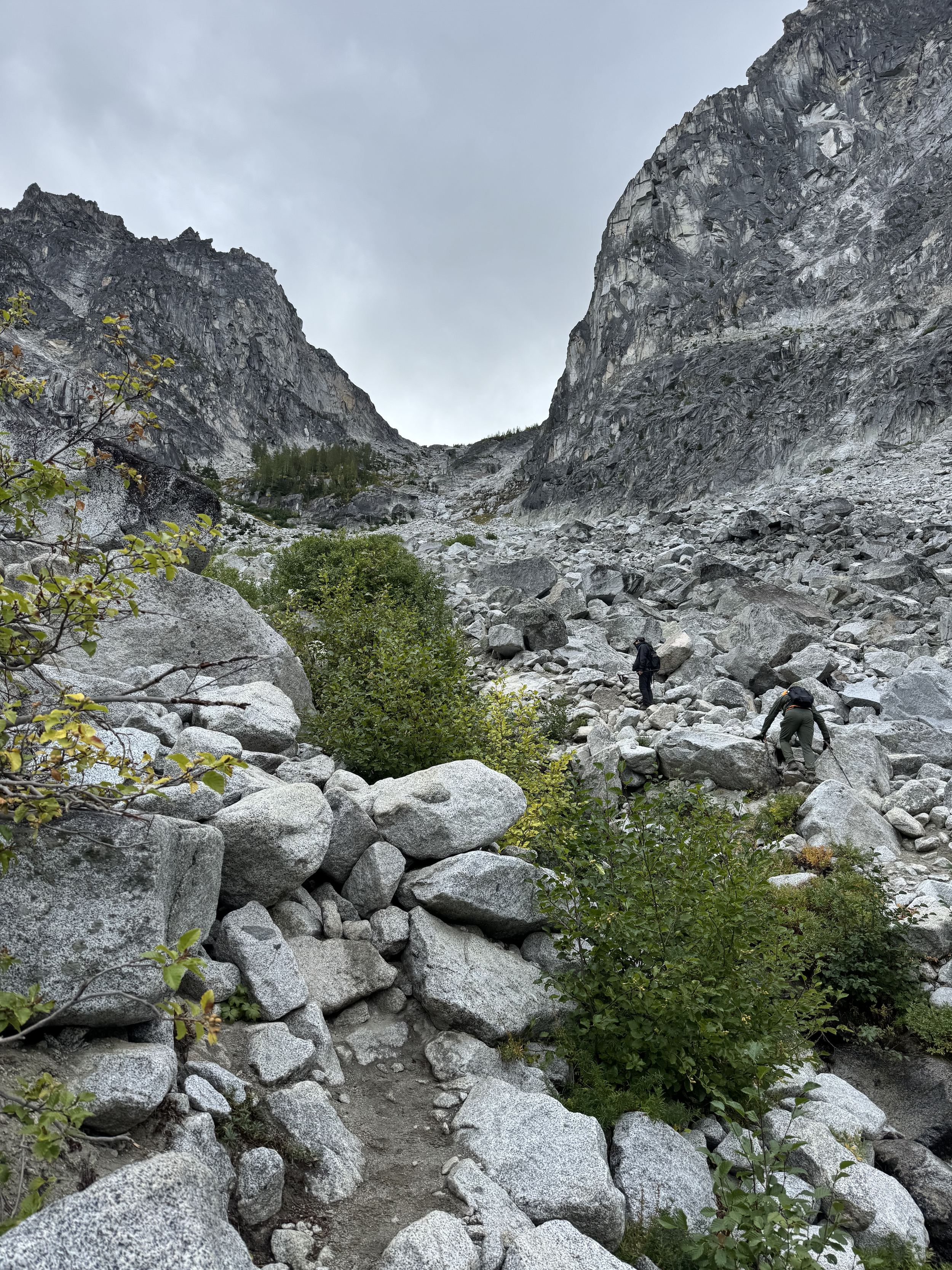 A person climbing over large gray rocks in a mountainous area with steep rocky cliffs on either side and a cloudy sky overhead.