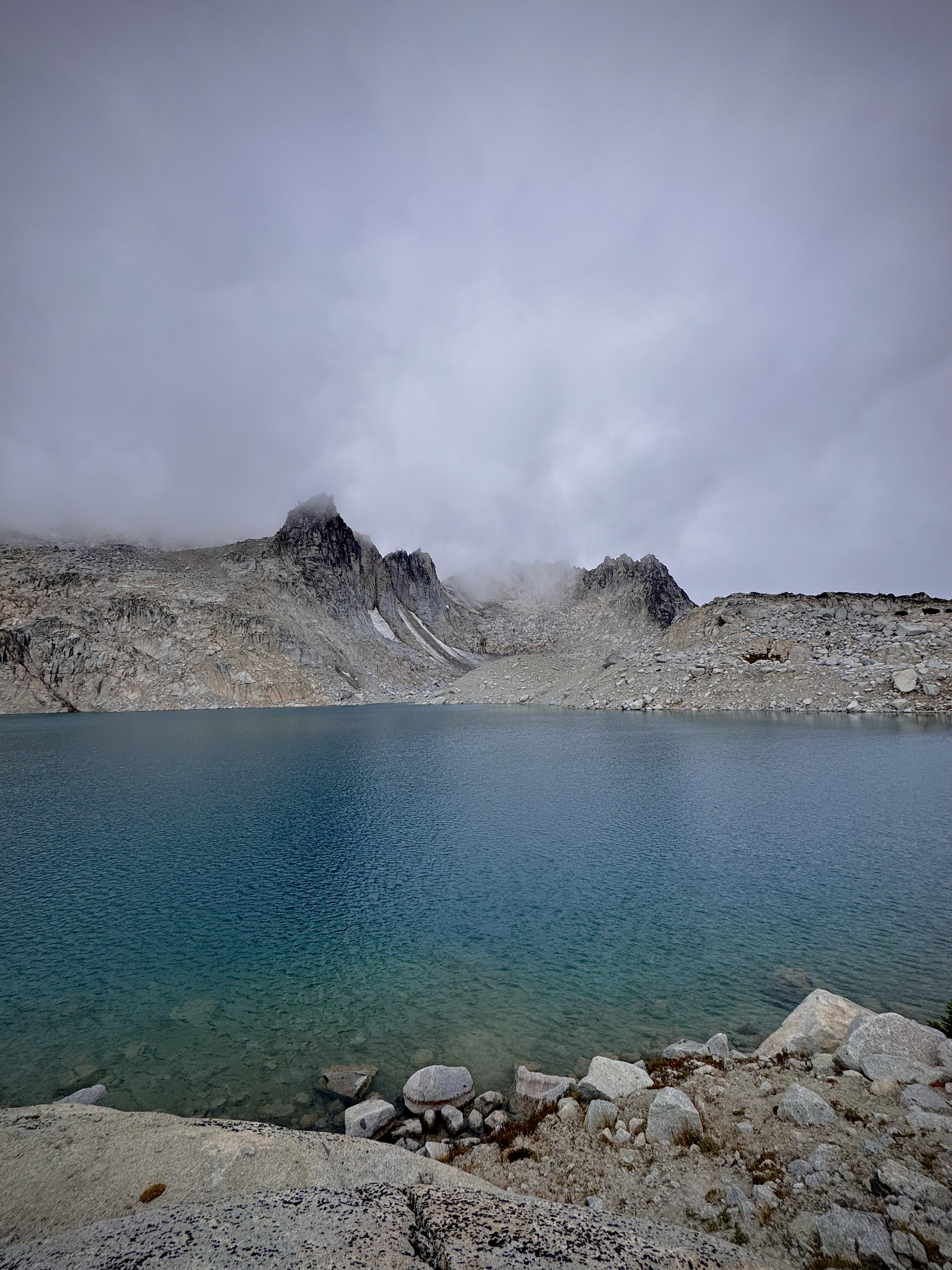 A mountain lake with clear blue water, rocky shoreline, rugged mountain peaks partially obscured by fog, and a cloudy sky.