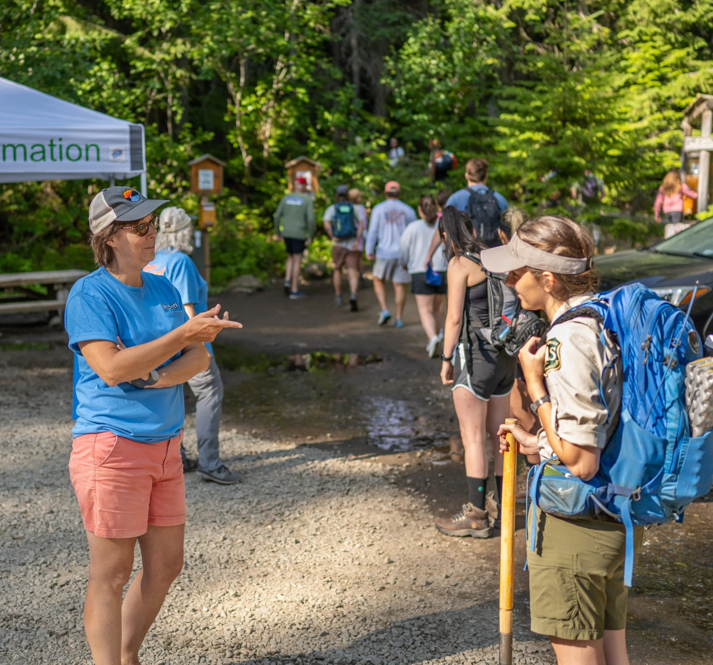 A group of people gathering outdoors near a wooded area, some carrying backpacks, with a woman in a blue shirt and cap engaged in conversation in the foreground, and informational signs and small structures in the background.