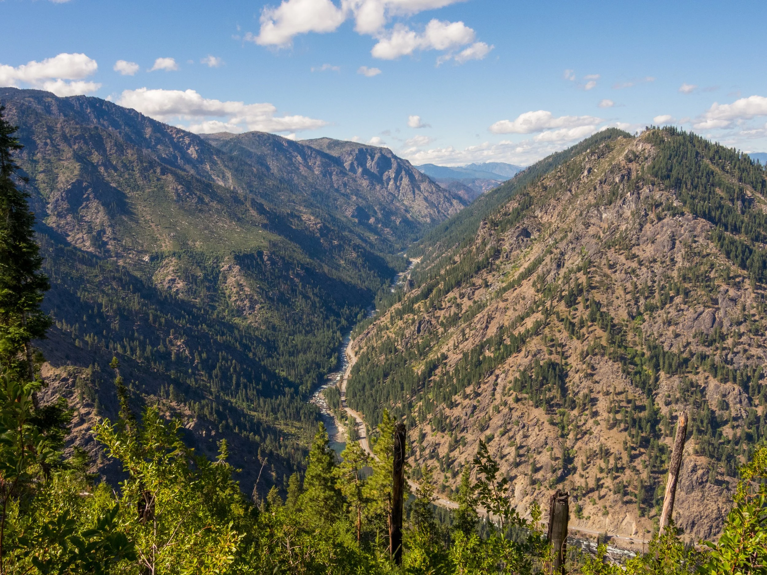 View of a mountainous valley with a winding river, forested slopes, and a partly cloudy sky.
