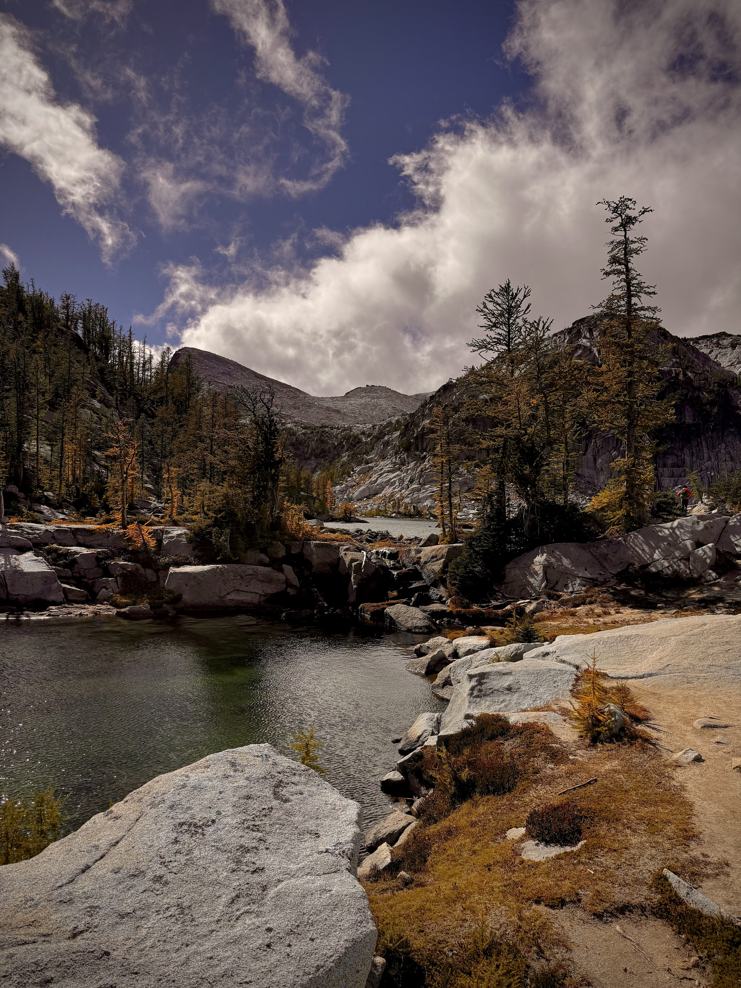 A mountain landscape with a river running through a rocky terrain, surrounded by trees with yellow and green foliage, under a sky with clouds and patches of blue.