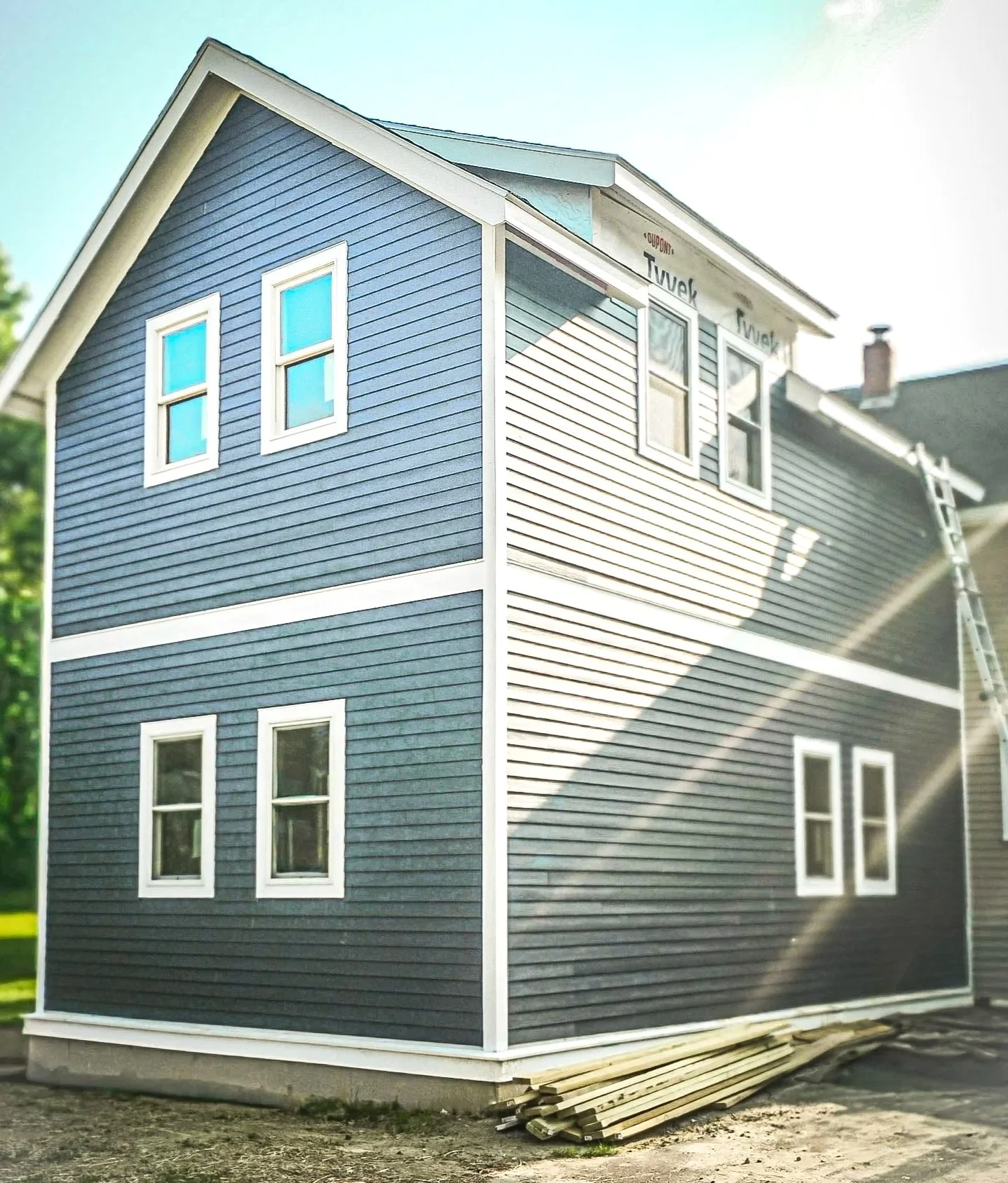 A two-story house under construction with dark blue siding, white window frames, and some windows installed. A ladder rests against the side, and wooden planks are on the ground near the house.