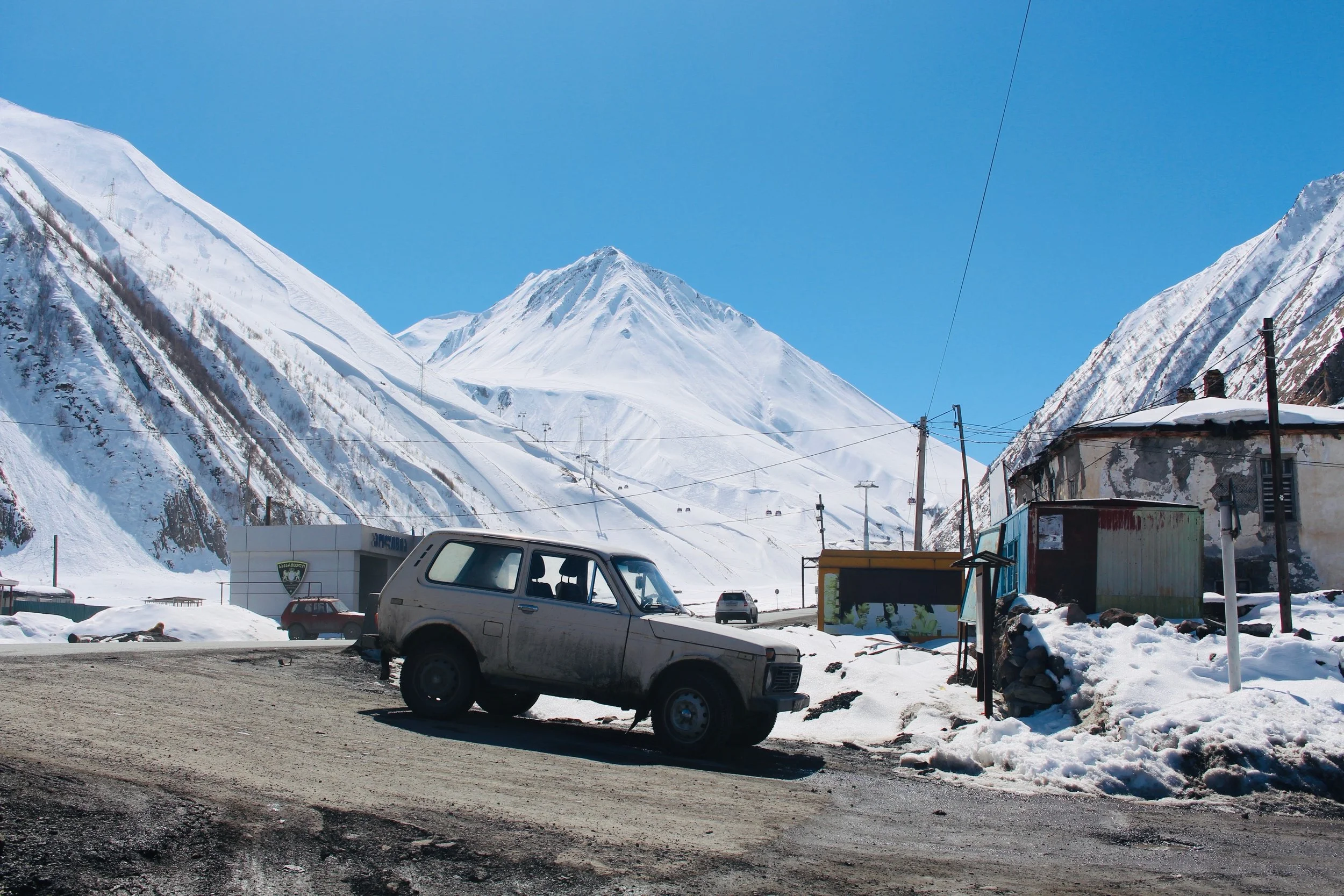 Snow-covered mountains against a bright blue sky with a small parking lot and a few parked cars in the foreground, including an old silver vehicle.