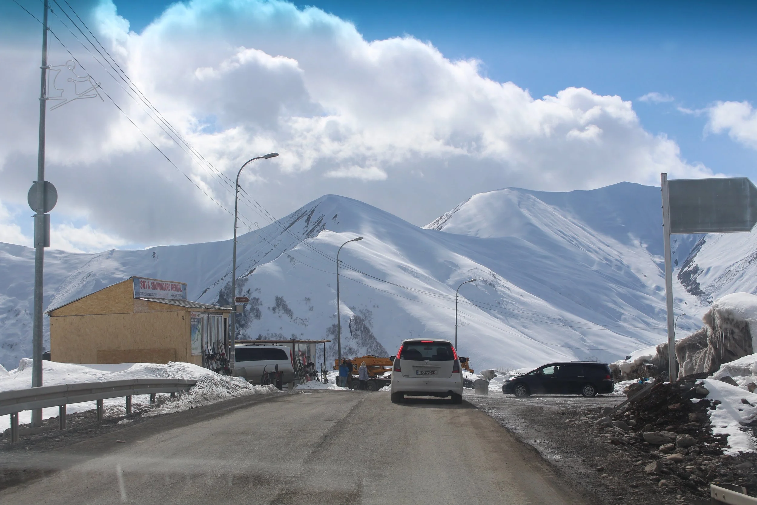 Snow-covered mountains under a partly cloudy sky with a ski and snowboard rental shop, parked vehicles, and ski equipment along the roadside.