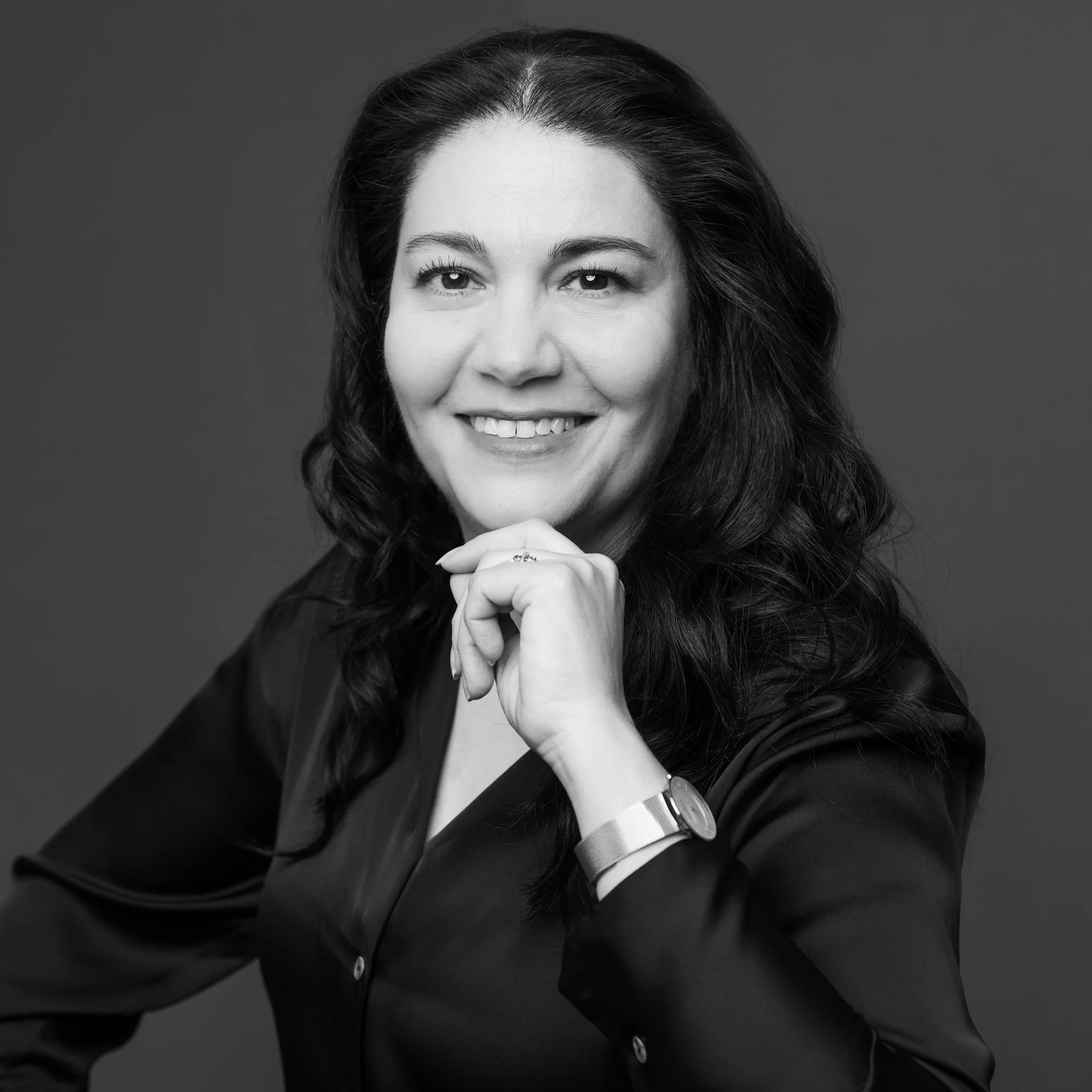 Portrait en noir et blanc d'une femme avec de longs cheveux bouclés, souriante, portant une montre et une tenue sombre, posant avec la main sous le menton contre un fond neutre.