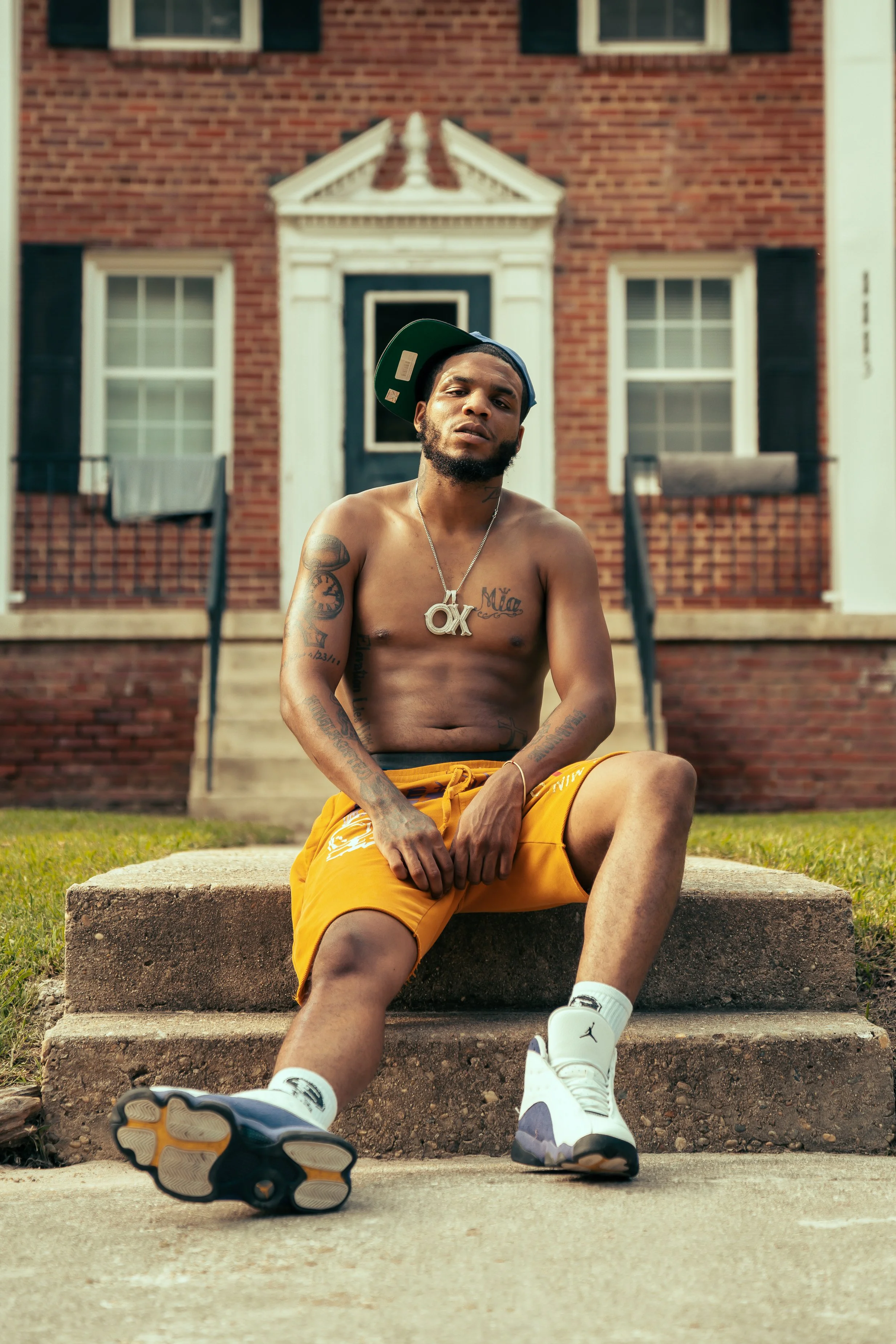 A shirtless young man with tattoos, wearing orange shorts, white sneakers, and a baseball cap, sitting on concrete steps in front of a red brick house.