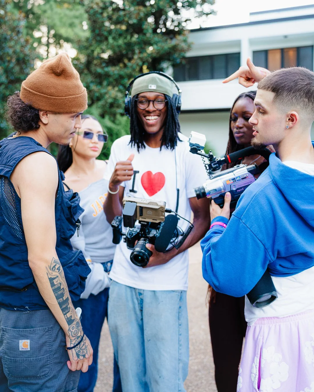 Group of young people, including a woman with tattoos and a beanie, a woman in sunglasses, and a man with a camera, gathered outside and engaged in a discussion, with one person pointing at the camera.