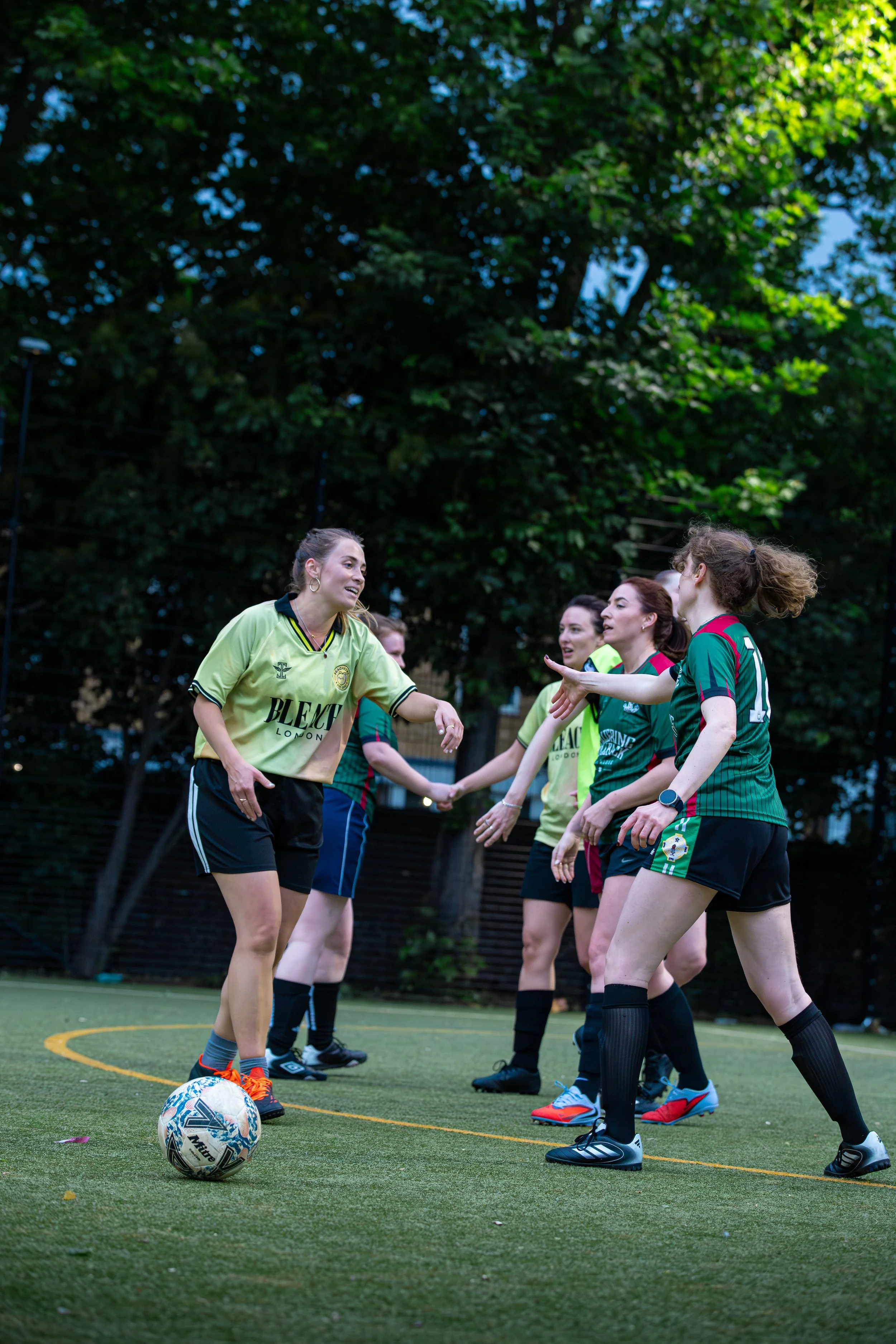 A group of women playing soccer on an outdoor field during daytime, with trees in the background, some women greeting each other.
