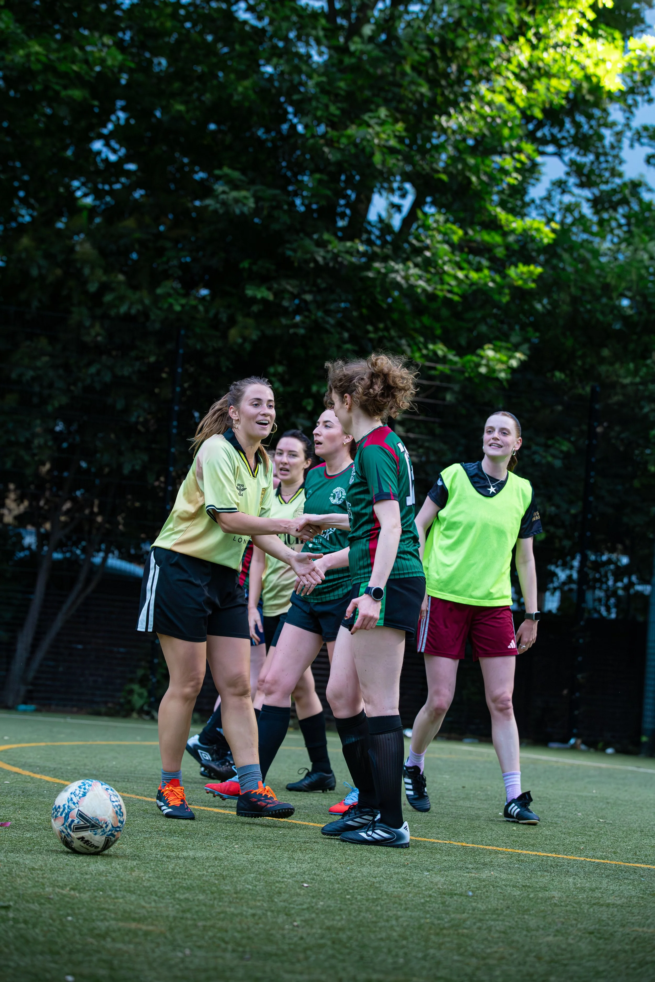 Women soccer players shaking hands after a match on an outdoor field with a green fence and trees in the background.