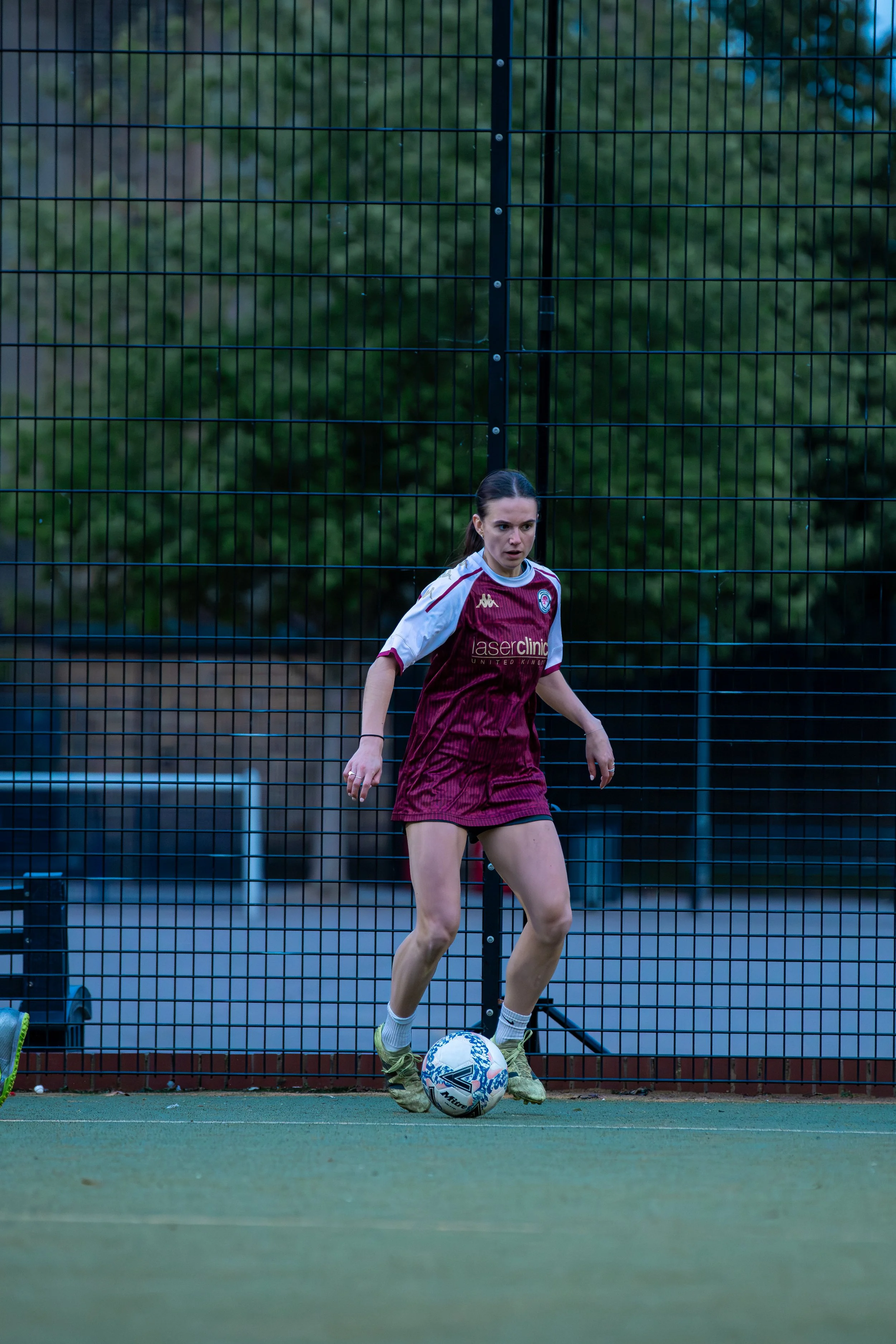 A woman in a maroon and white soccer uniform kicks a ball on a field, standing behind a tall black metal fence with trees in the background.