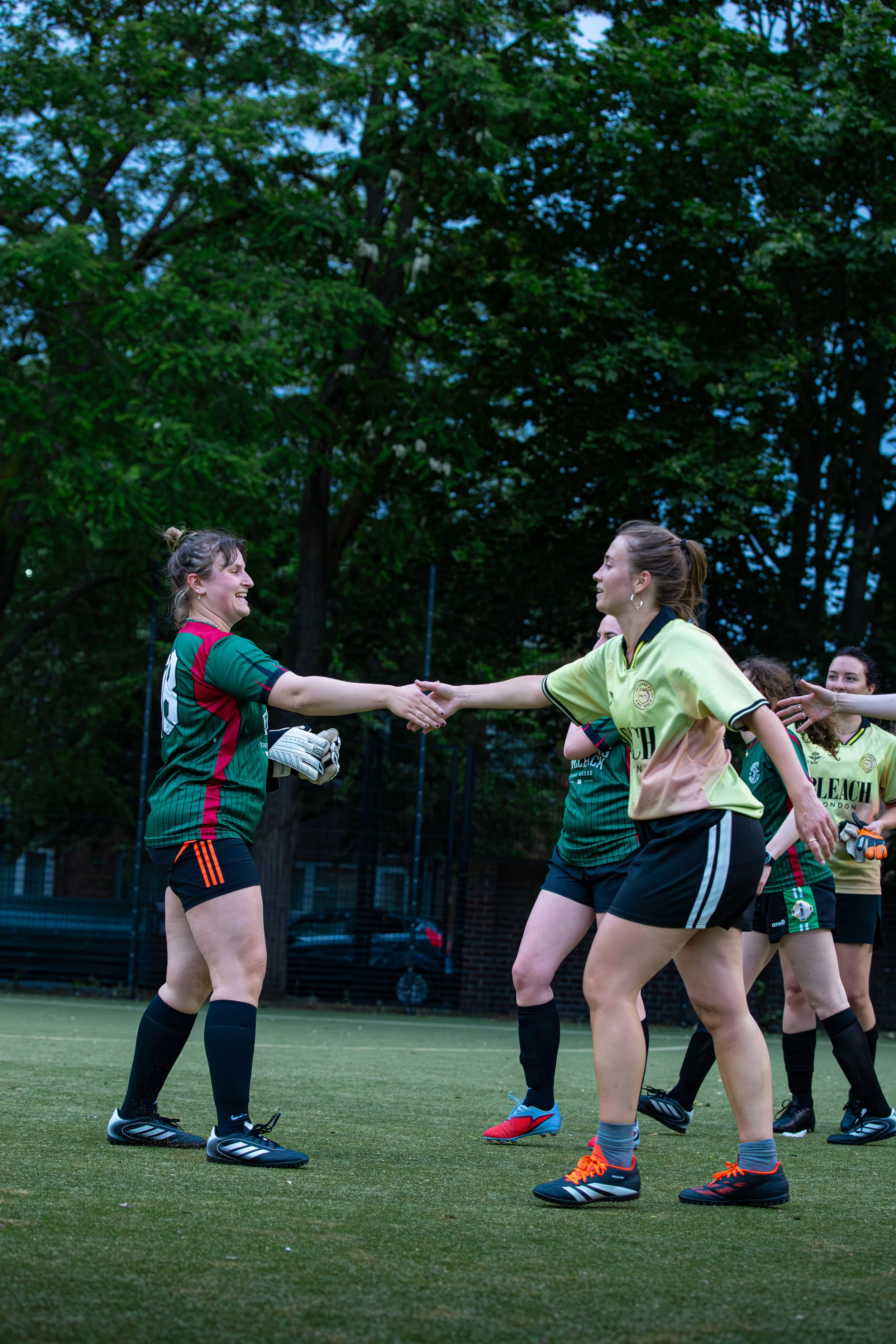 A group of female soccer players on the field, shaking hands after a game, with trees in the background.