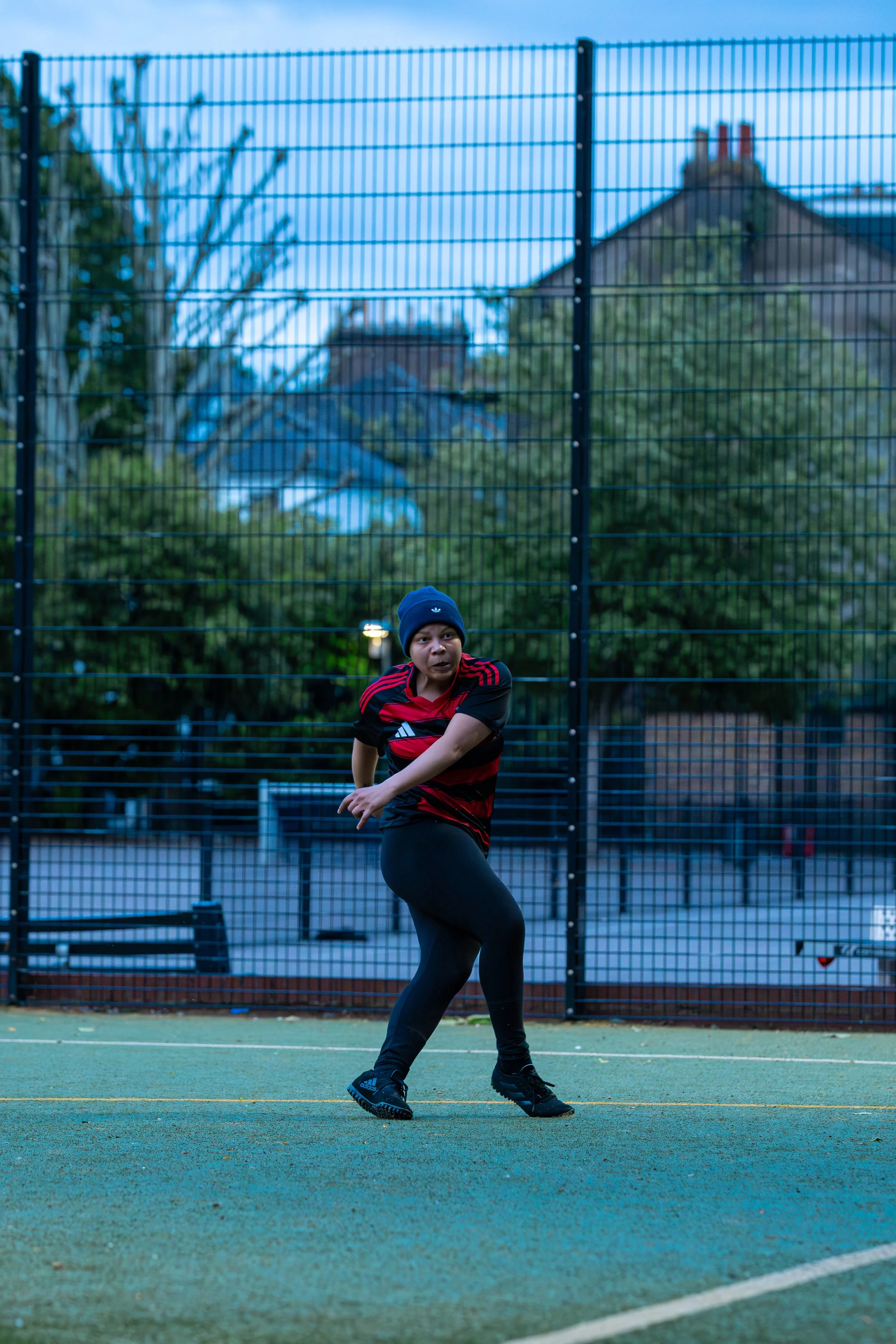 Young woman playing tennis on an outdoor court, wearing a blue beanie, red and black sports shirt, black leggings, and tennis shoes, with a tall fence and houses in the background.