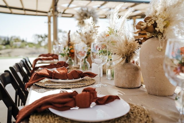 A long dining table set with white plates, rust-colored cloth napkins, clear wine glasses, and large floral arrangements in beige vases, under a canopy with outdoor scenery in the background.