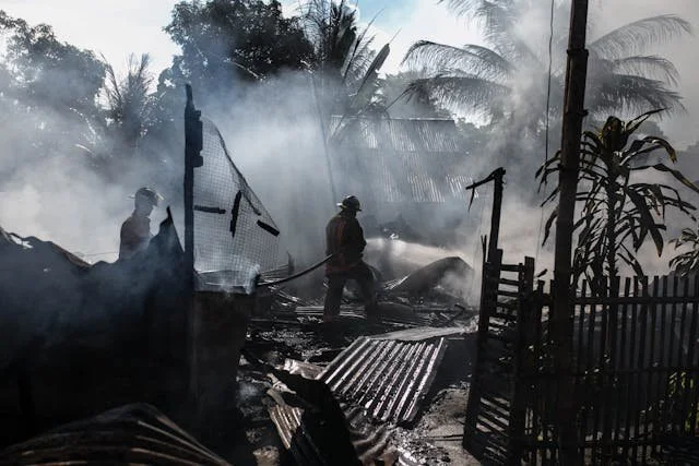 Firefighters spraying water on a smoldering area of a burned-down structure surrounded by smoke, with palm trees and a fence in the background.