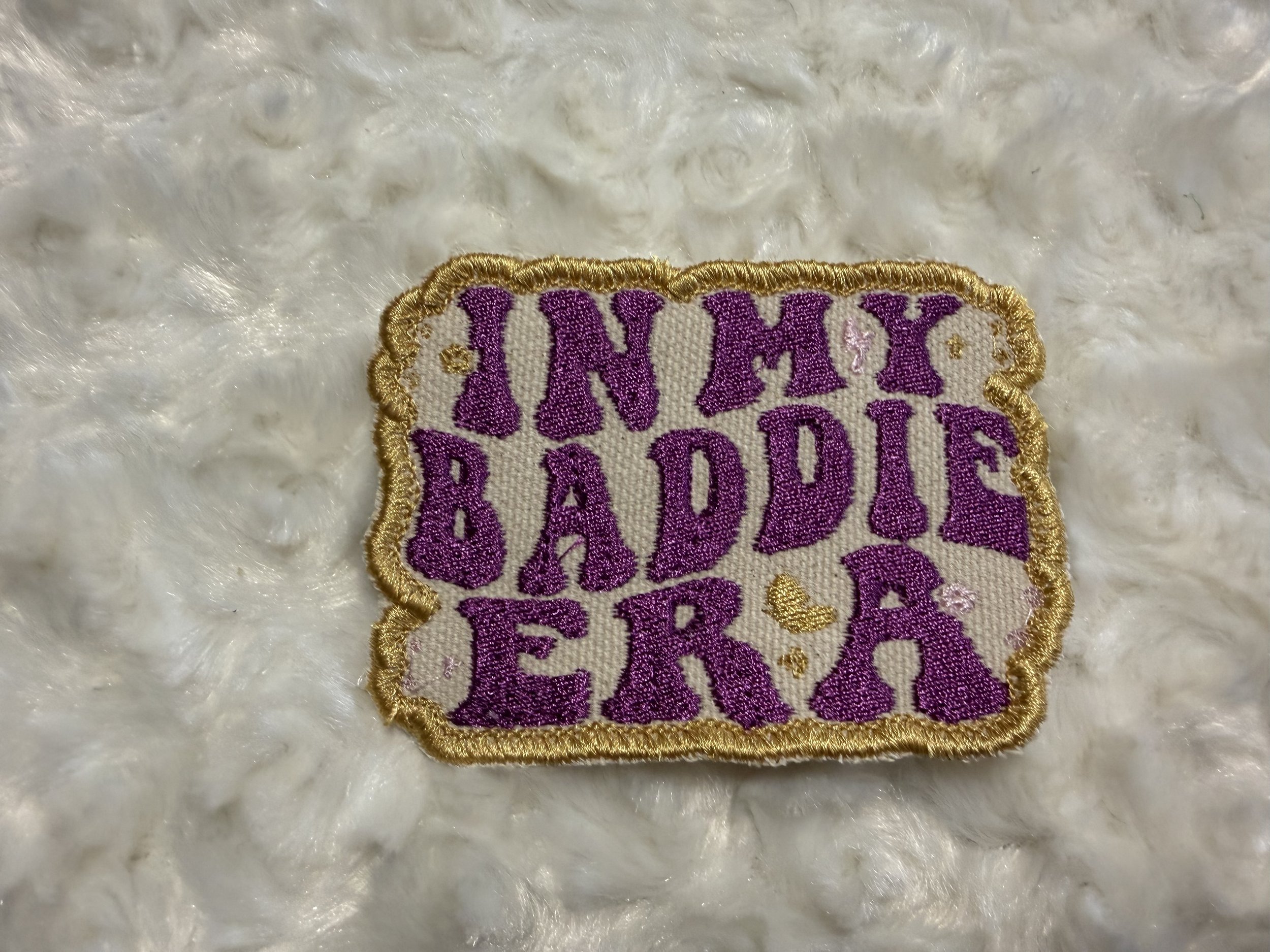 Close-up of a embroidered patch with purple text and gold border on a white furry surface.