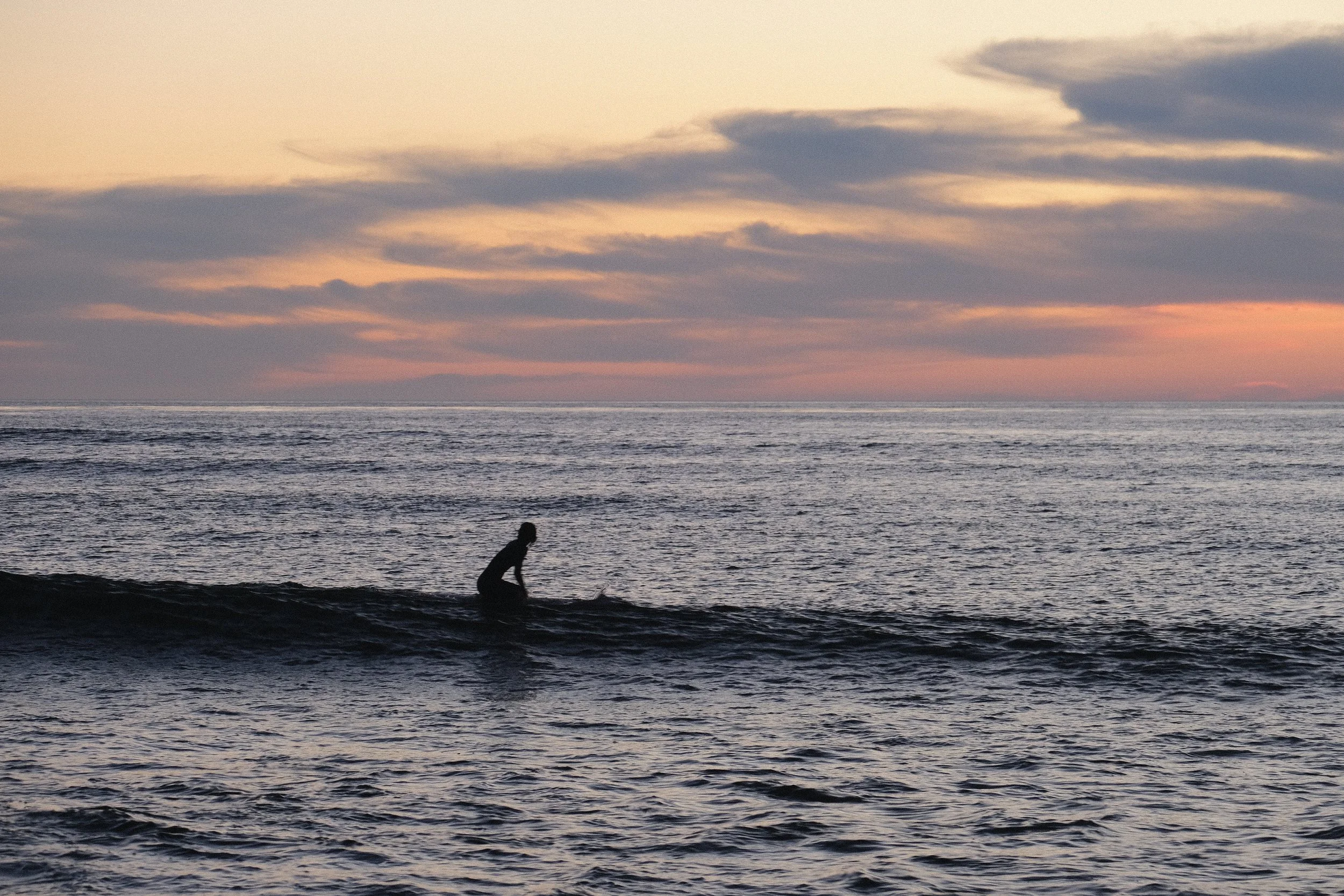A person is kneeling on a surfboard in the ocean, with a colorful sky and clouds at sunset in the background.
