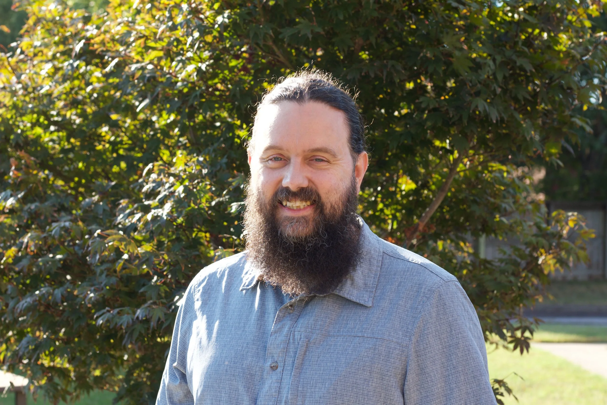 A man with a long beard and mustache smiling outdoors with sunlight shining on his face, standing in front of green leafy trees.