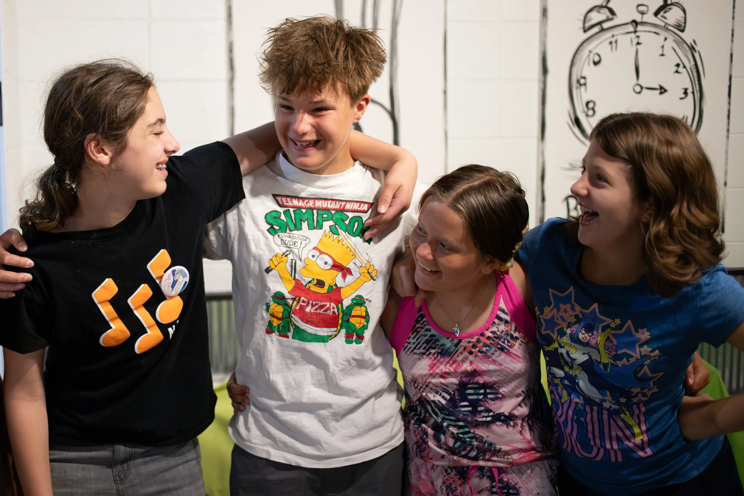 Group of five children hugging and laughing together indoors, with a large clock drawing on the white wall in the background.