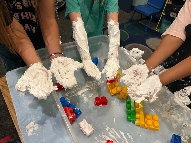Children's hands covered in white foam playing with large colorful building blocks on a table in a classroom or play area.