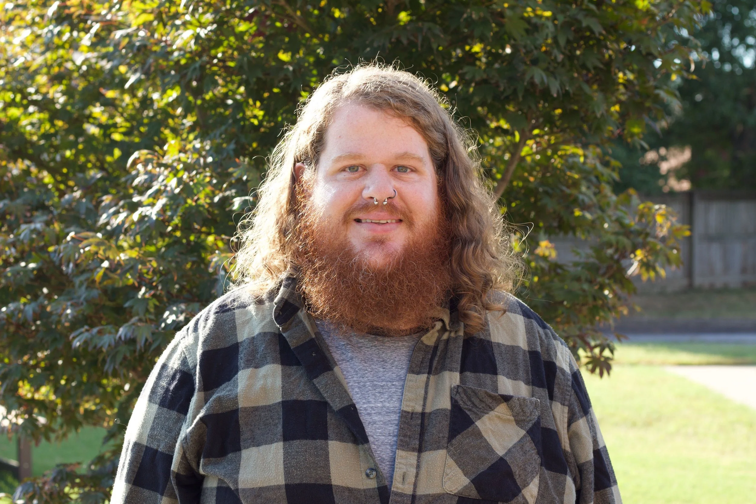 A young man with long curly hair, a beard, and septum piercings smiling outdoors, wearing a checkered flannel shirt.