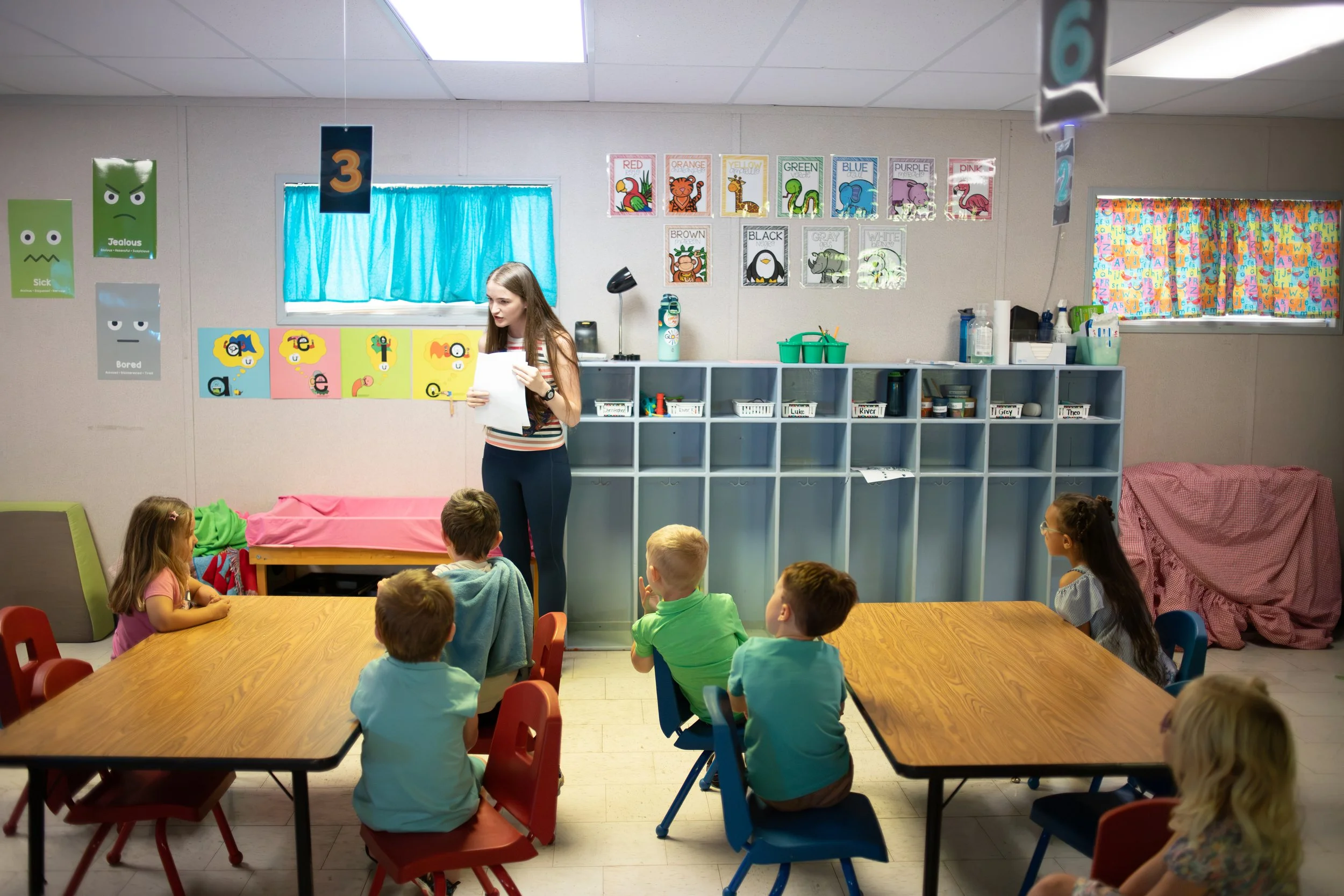 A classroom with young children sitting at a table, listening to a female teacher who is holding papers. The classroom has colorful posters and decorations, including educational posters on the wall, and a blue curtain over the window.