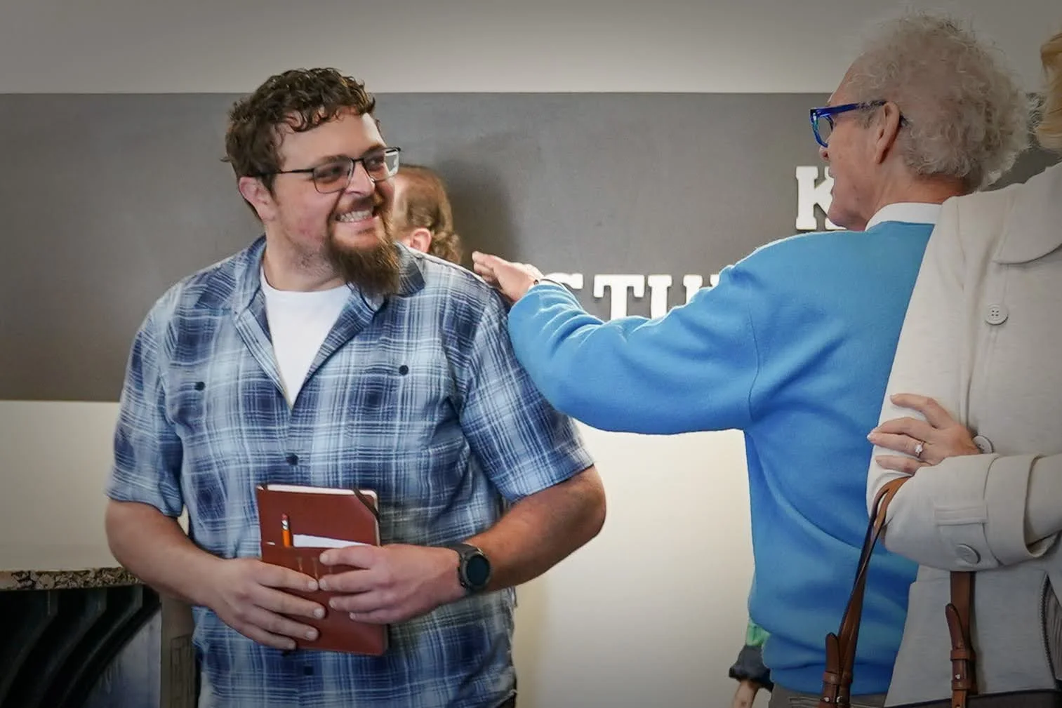 A young man with glasses and a beard smiling as an older woman with curly gray hair and glasses pats his shoulder, standing in an indoor setting.