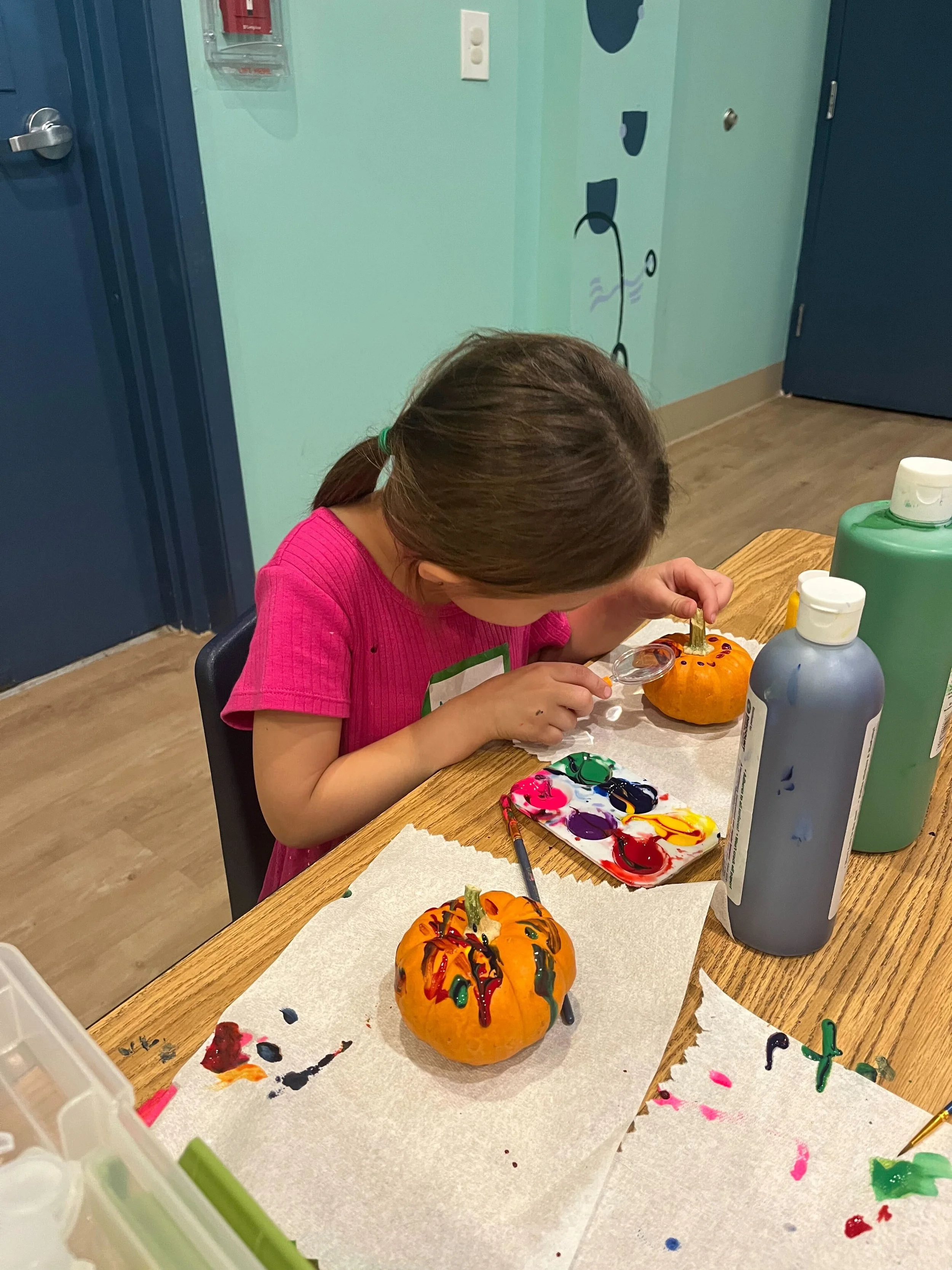 A young girl with brown hair in a ponytail, wearing a pink shirt, is painting a small pumpkin with colorful decorations at a table. The table has paint supplies, and the pumpkin has bright colors and patterns. In the background, there is a painted wall with a whimsical black line drawing of a vine with leaves.