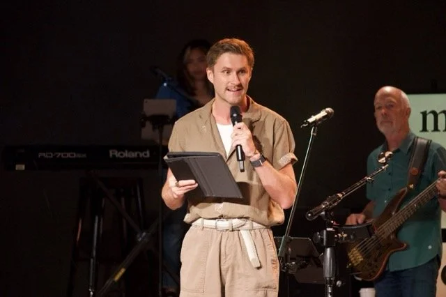 A man holding a microphone and a tablet on stage during a performance or presentation, with musicians playing instruments in the background.