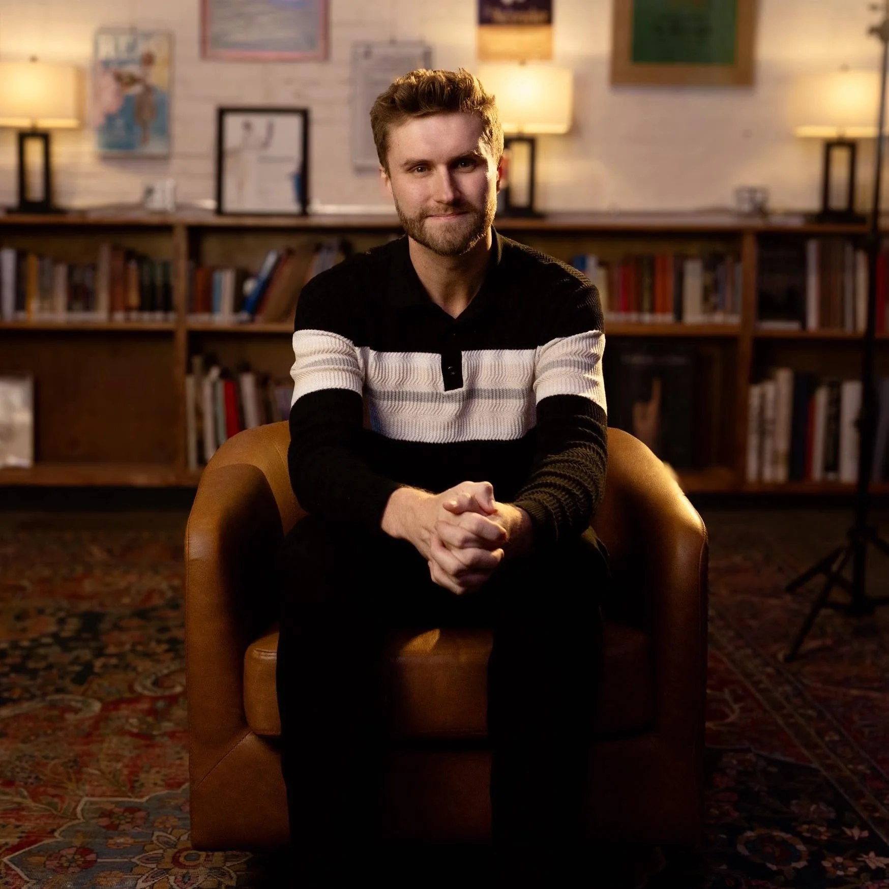 A man with short light brown hair and a beard sitting on a yellow armchair in a warmly lit room, with bookshelves and framed artwork on the wall behind him.