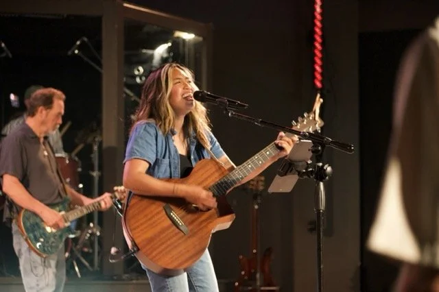 A female singer playing an acoustic guitar and singing into a microphone on stage, with a male guitarist playing electric guitar in the background.