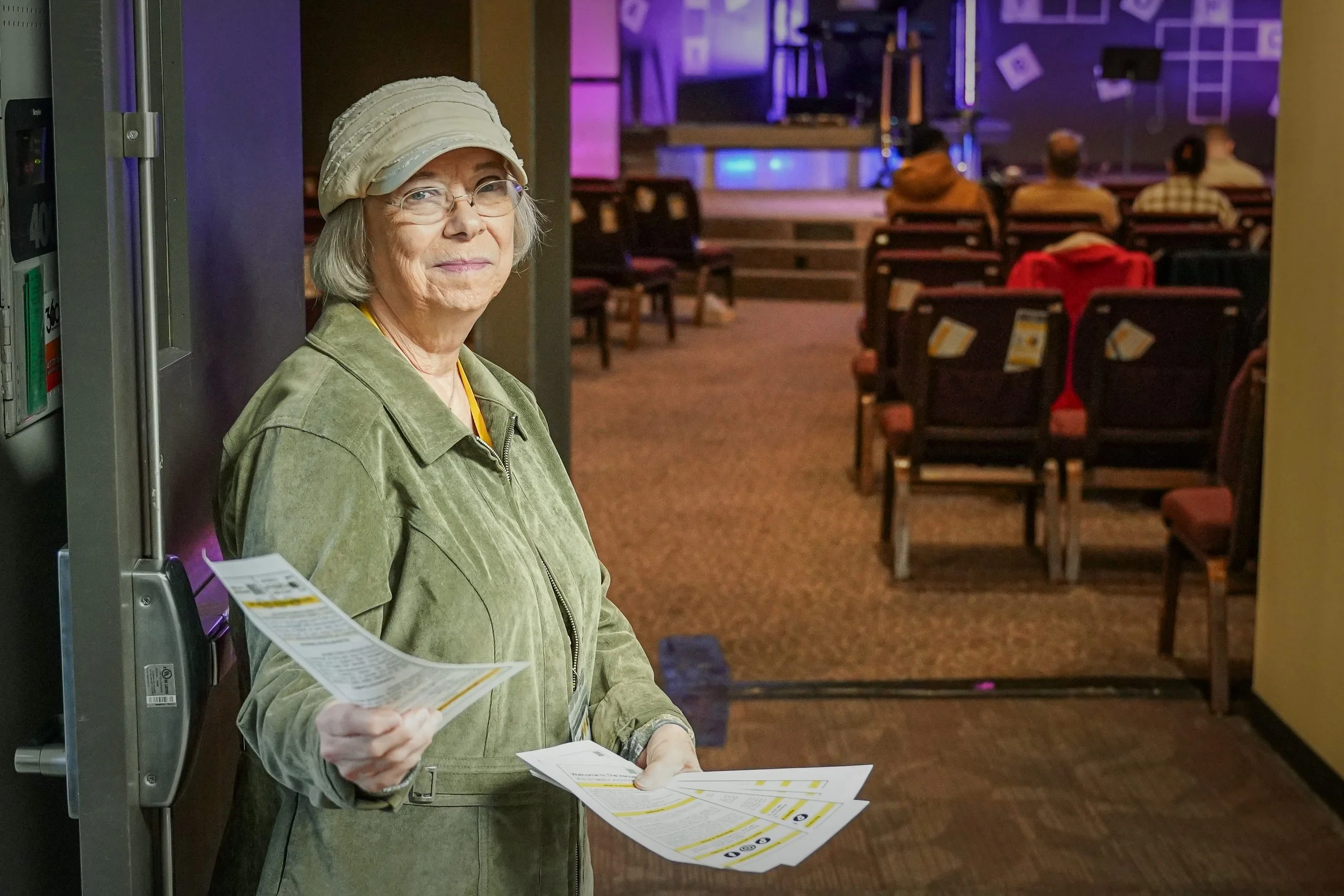 An elderly woman with glasses, wearing a beige hat and green jacket, holds flyers or brochures and stands near an elevator in a dimly lit auditorium or event space. There are several chairs and people in the background, and the stage is set with purple lighting and abstract decorations.