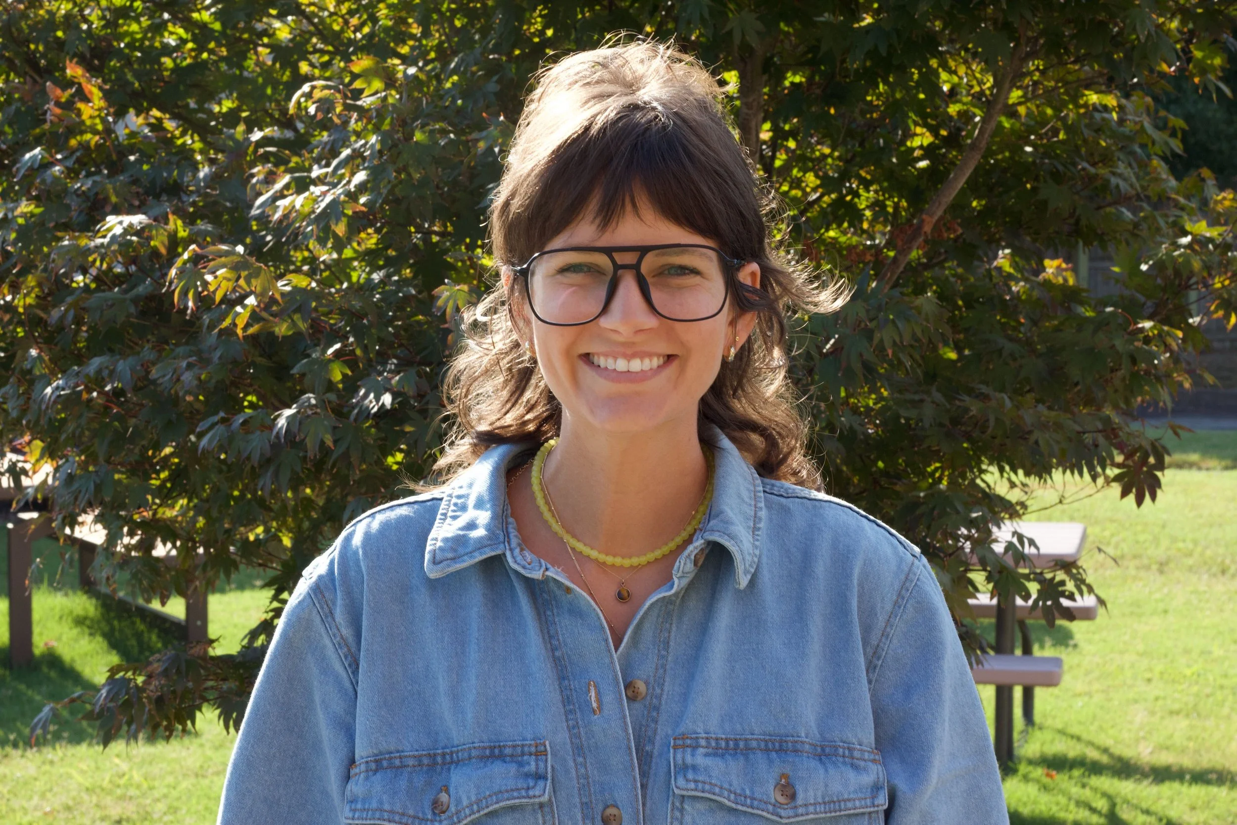 A woman with curly brown hair wearing glasses, a denim jacket, and multiple necklaces, smiling outdoors in front of a bush with green leaves.
