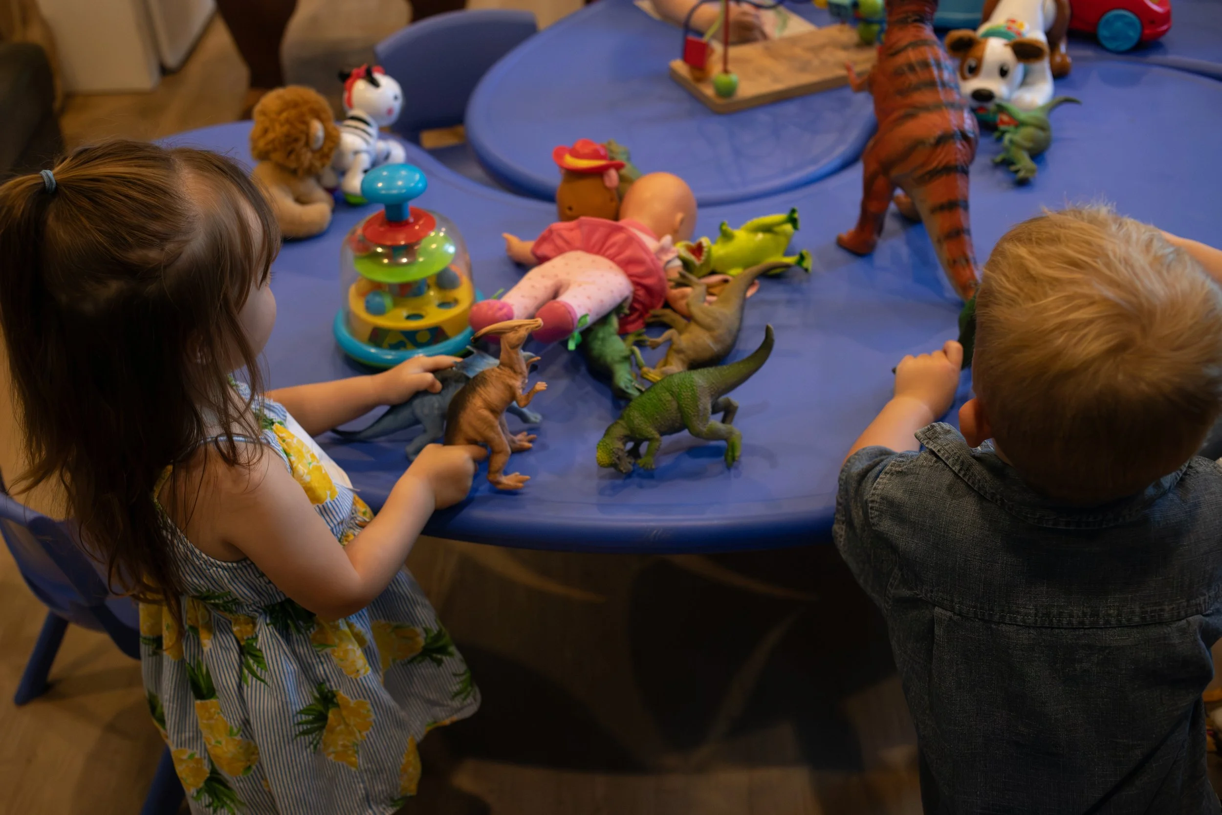 Children sitting around a blue table with various toys, including stuffed animals and dinosaur figures, during a playtime activity.