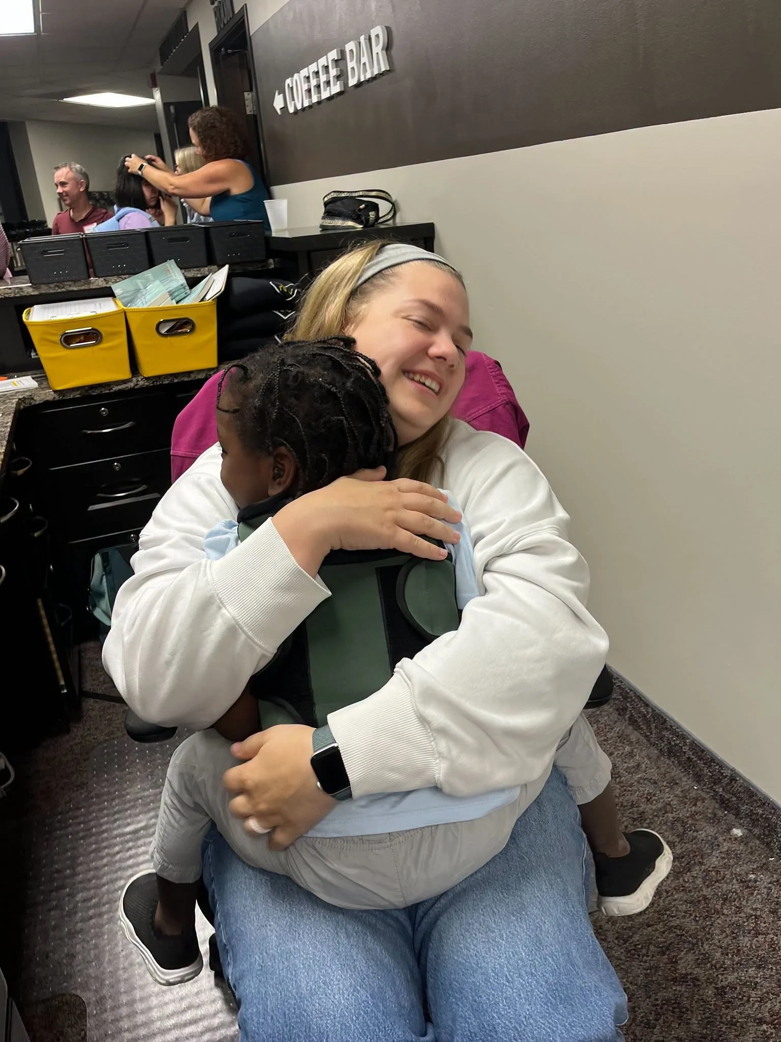 A woman with curly hair and a gray headband hugging a young girl with braided hair and a light blue hoodie in a seated position in an indoor setting. Behind them, a coffee bar sign is visible on the wall, and there is a counter with yellow and black storage bins. In the background, a group of people are engaging in conversation near a door.