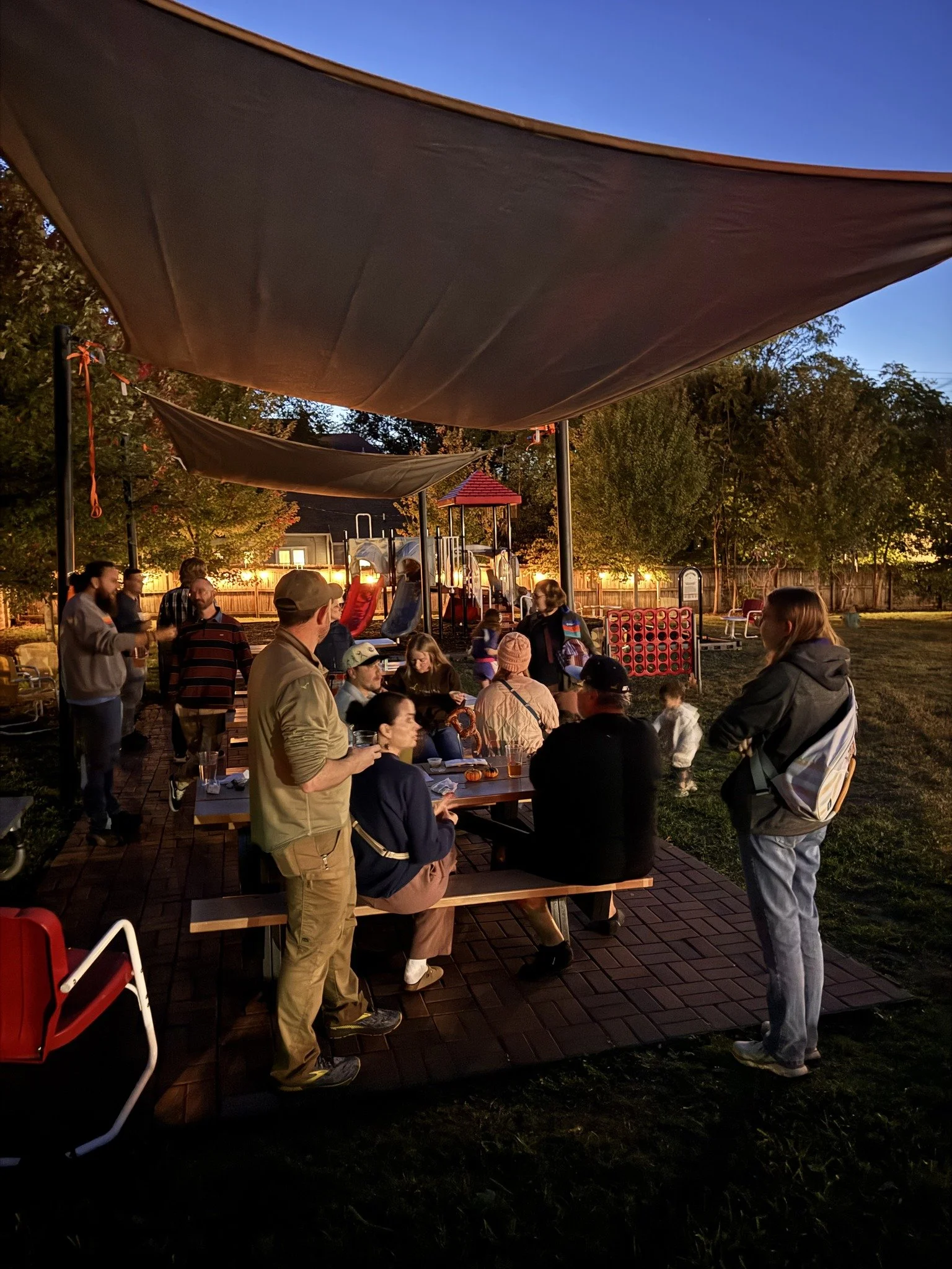 People gathered outdoors in a backyard at dusk, sitting and standing around picnic tables, with some standing and talking, overlooking a playground with slides and a red-roofed play structure, under a large canopy shade, surrounded by trees and a wooden fence.