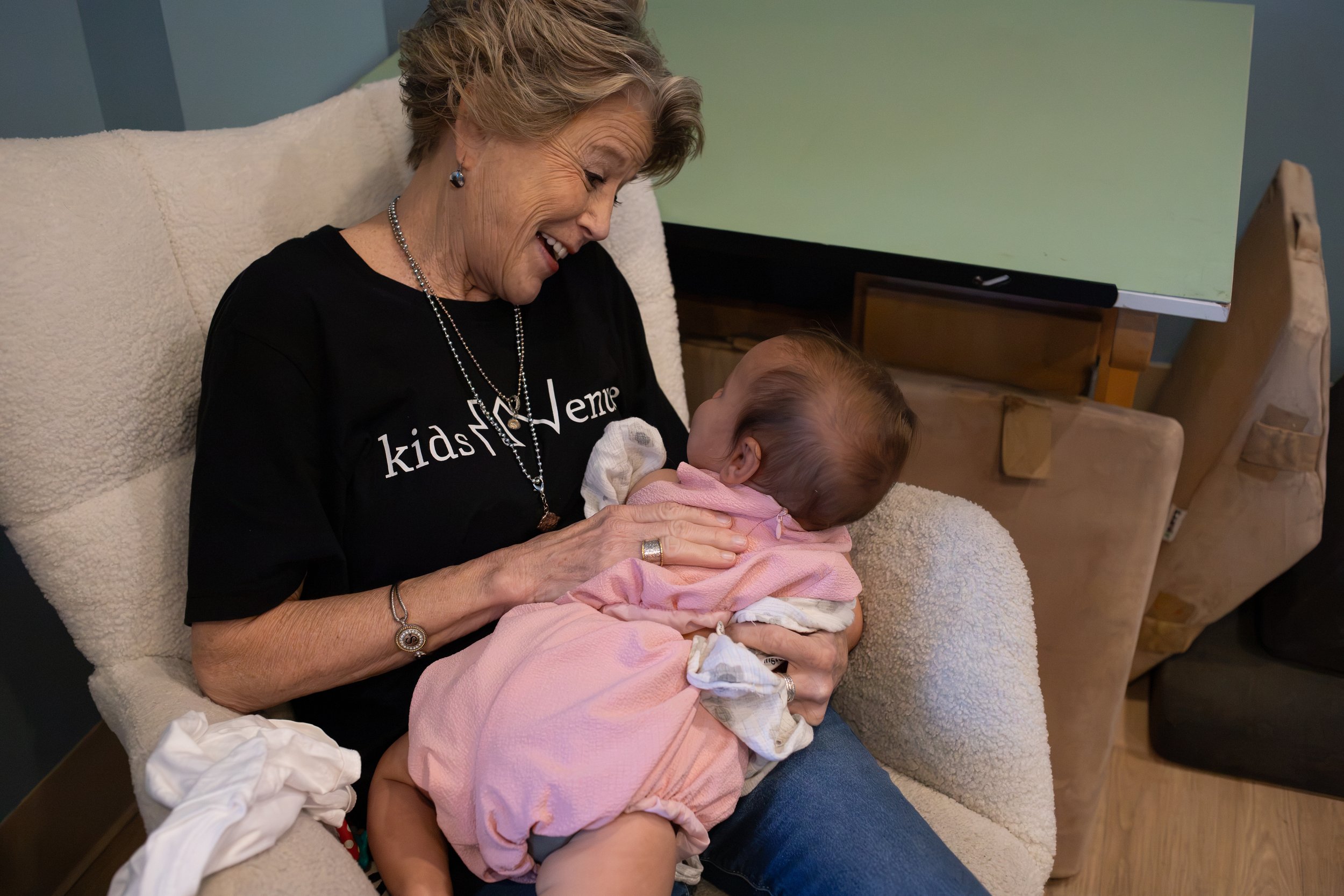 An elderly woman with short hair and jewelry, smiling while holding a baby girl dressed in pink, sitting on a cream-colored couch.