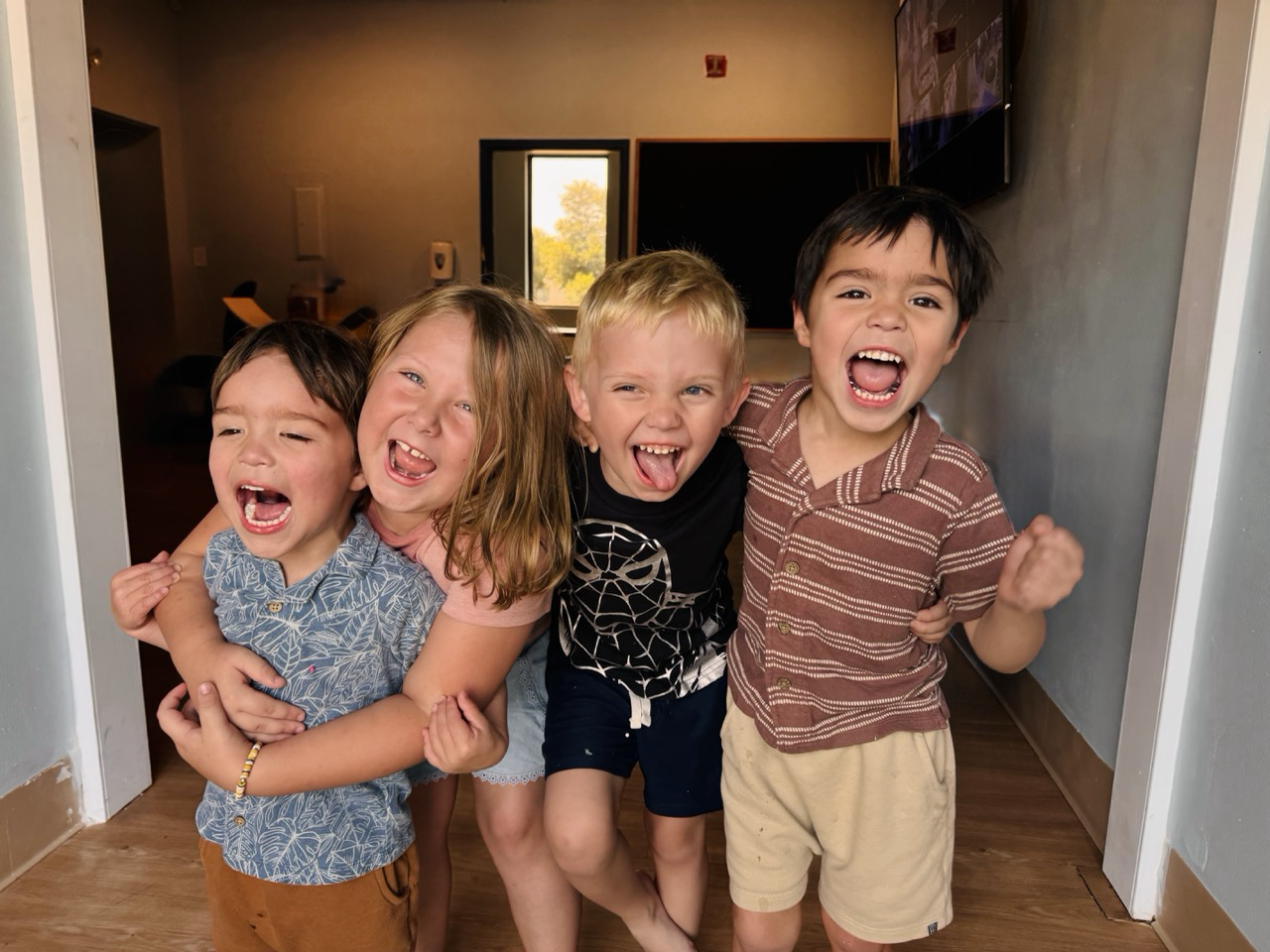 Four children are happily posing together indoors, smiling and laughing, with arms around each other.