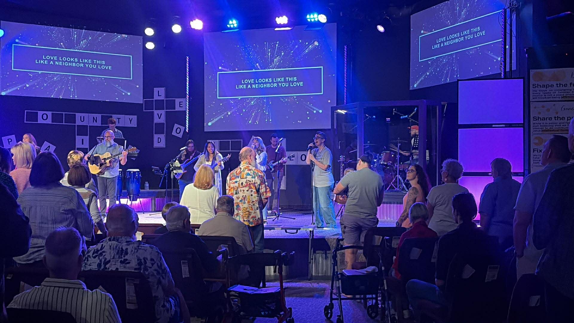 A church service or gathering with a live band performing on stage. Several people are standing and singing, while others are seated or standing in the audience. The stage has multiple screens displaying the lyrics, "Love looks like this like a neighbor you love," and various symbols and decorations. The lighting is blue and purple, creating a vibrant atmosphere.