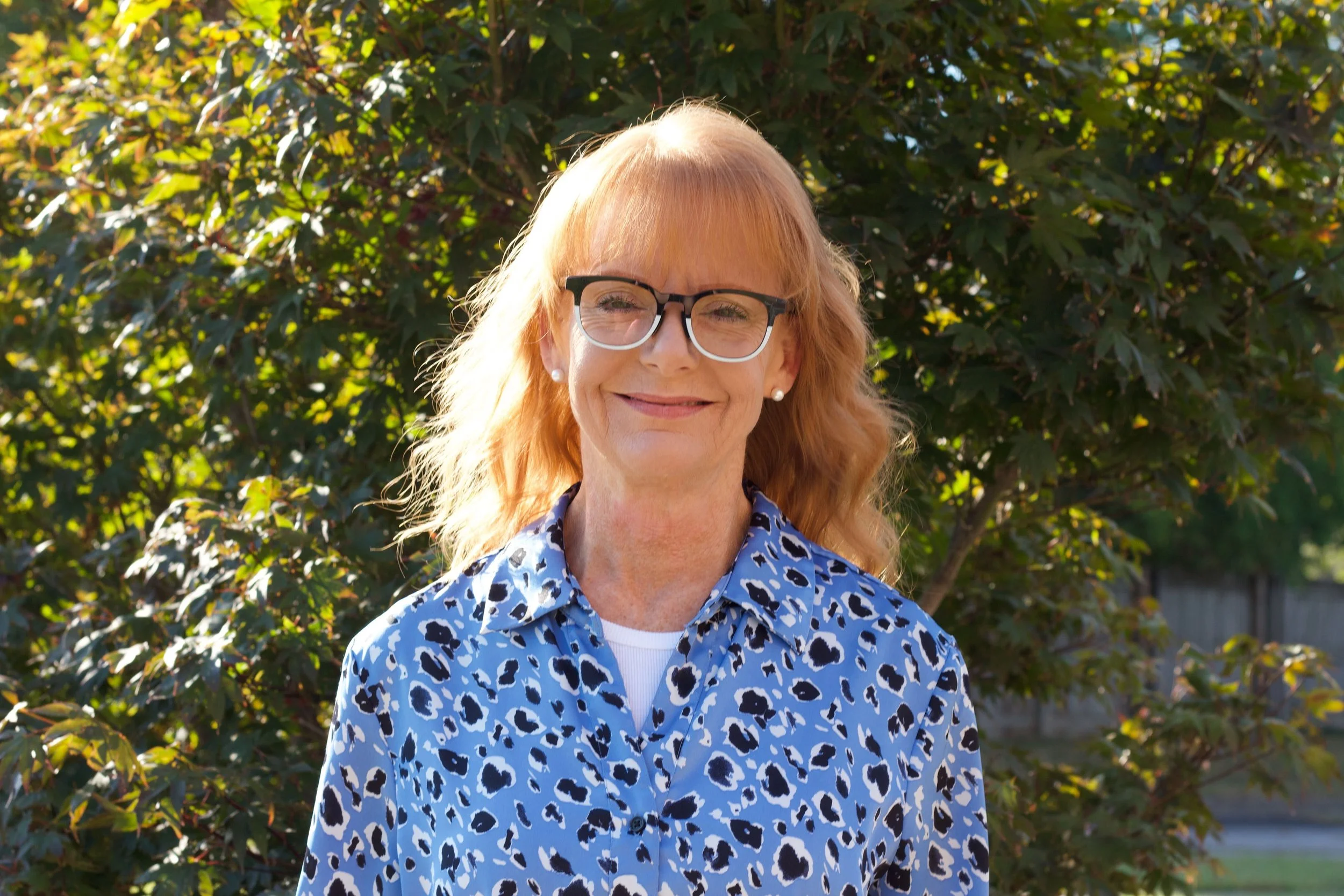 A smiling woman with blonde hair wearing glasses and a blue leopard print shirt standing outdoors in front of a leafy green bush.