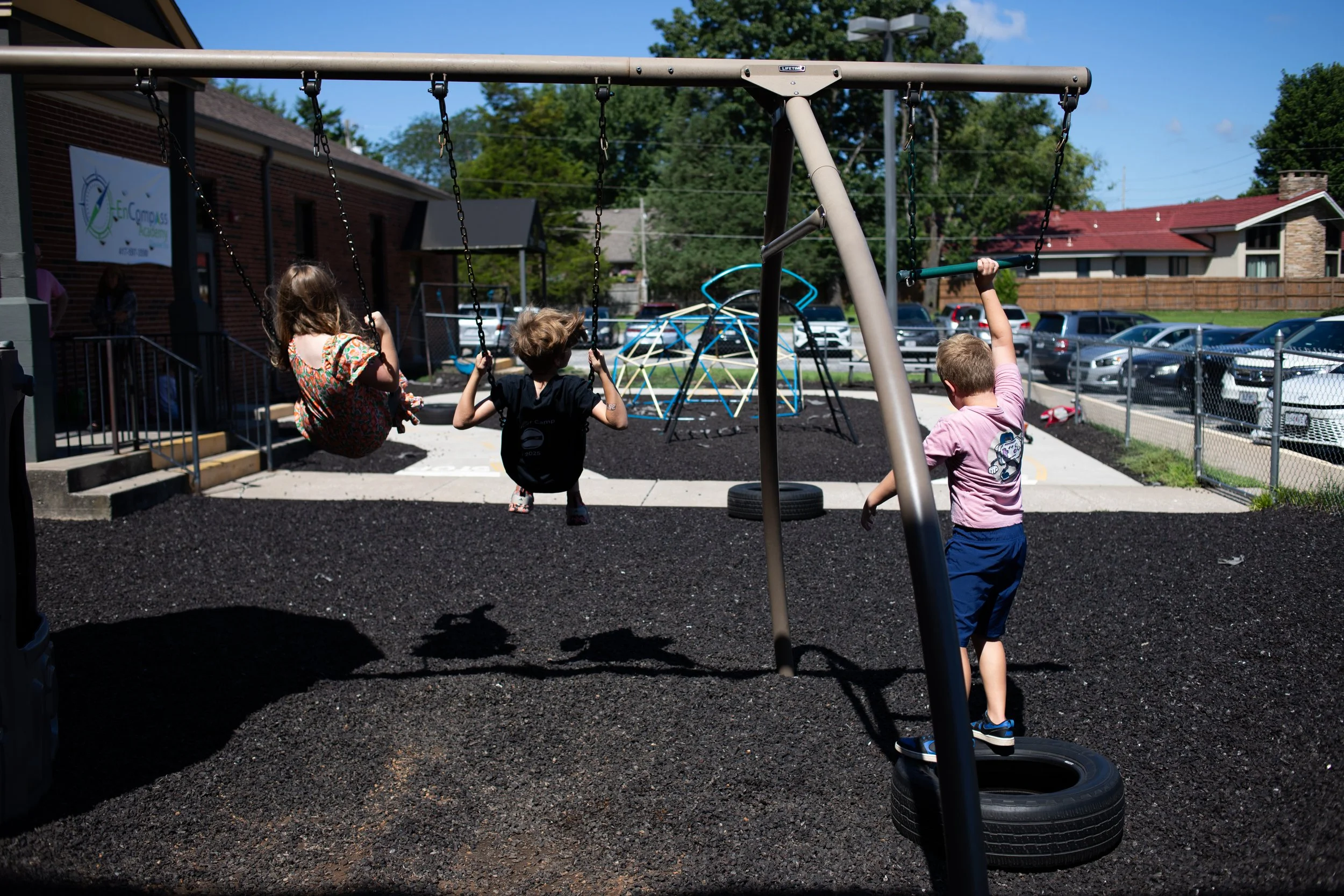 Children playing on a playground swings with a tire swing and climbing structure under a blue sky and green trees.