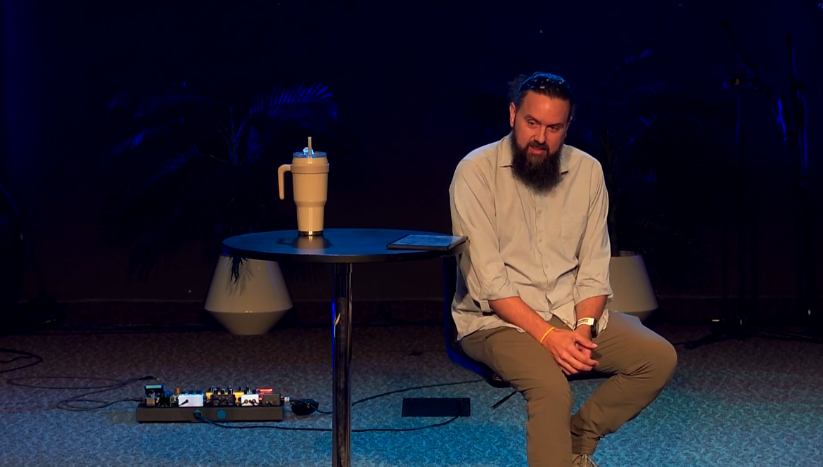 Man with a beard sitting on a chair in a dimly lit room with plants, a table with a drink, and a pedalboard on the floor.
