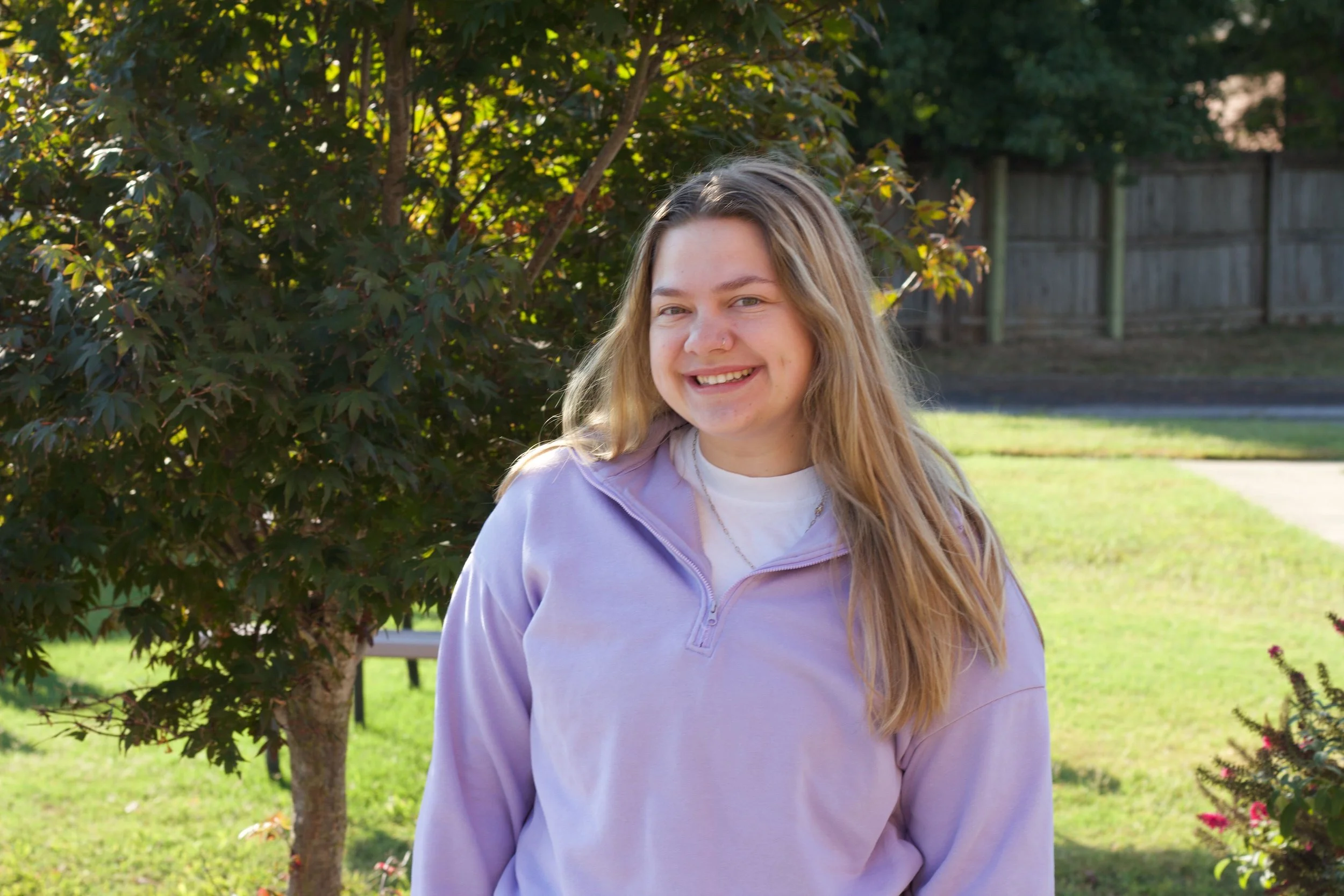 A young woman with long blonde hair smiling outdoors near a bush and a wooden fence.