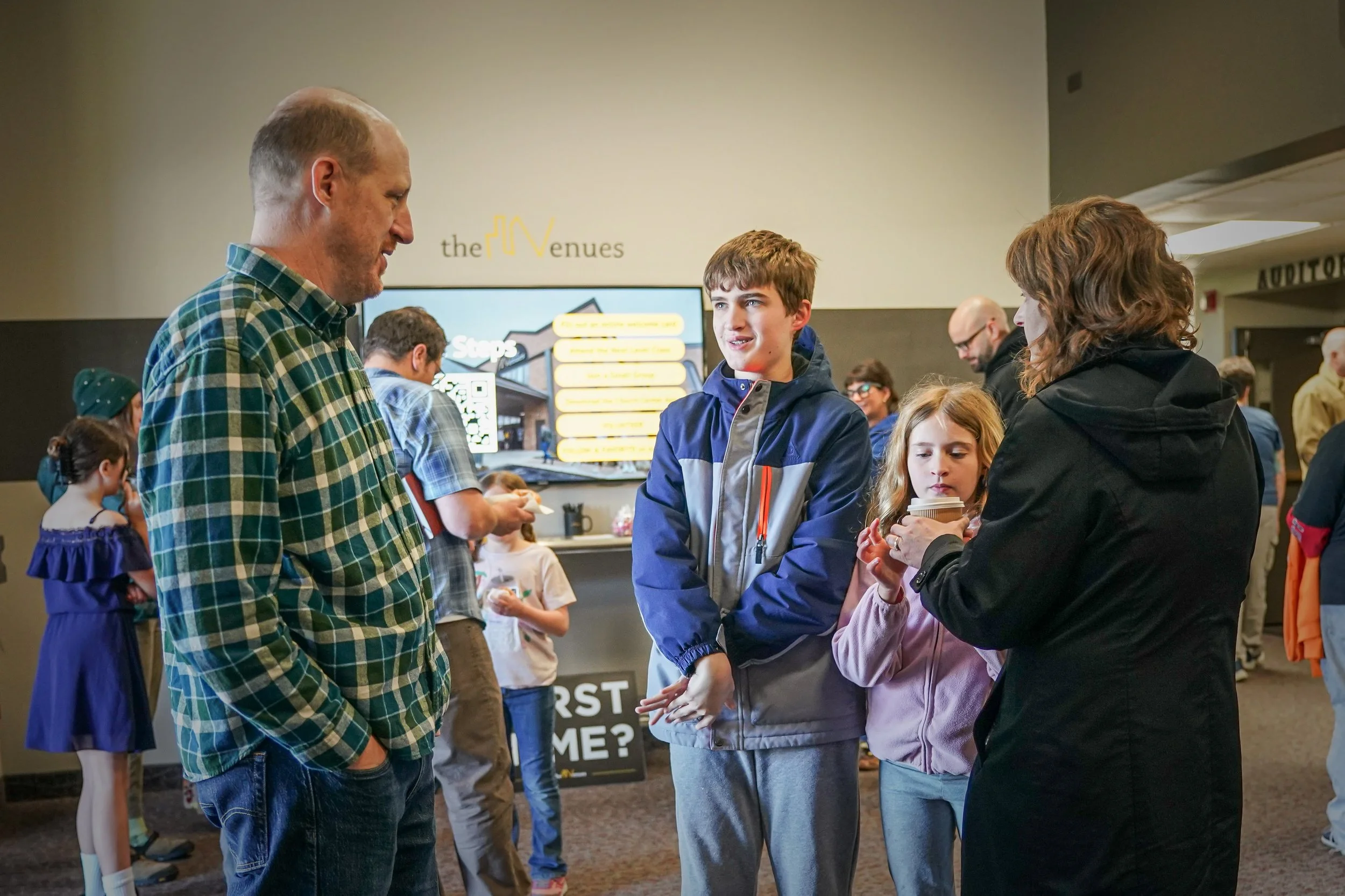 A man in a green plaid shirt and a woman in a black jacket are talking to two children, a boy and a girl, in a crowded indoor setting. In the background, several other people and a large digital screen displaying text are visible.