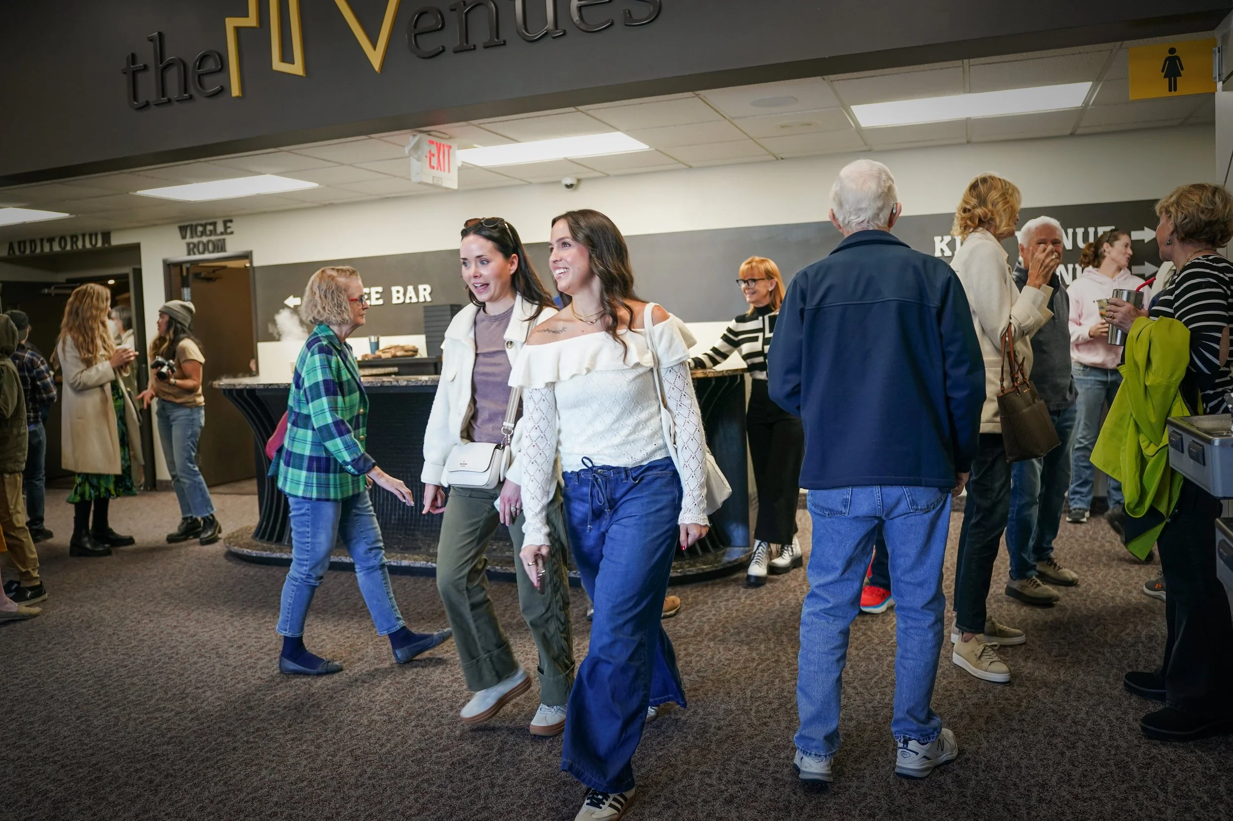 People walking and socializing in a theater lobby near a concession stand and restroom signs.