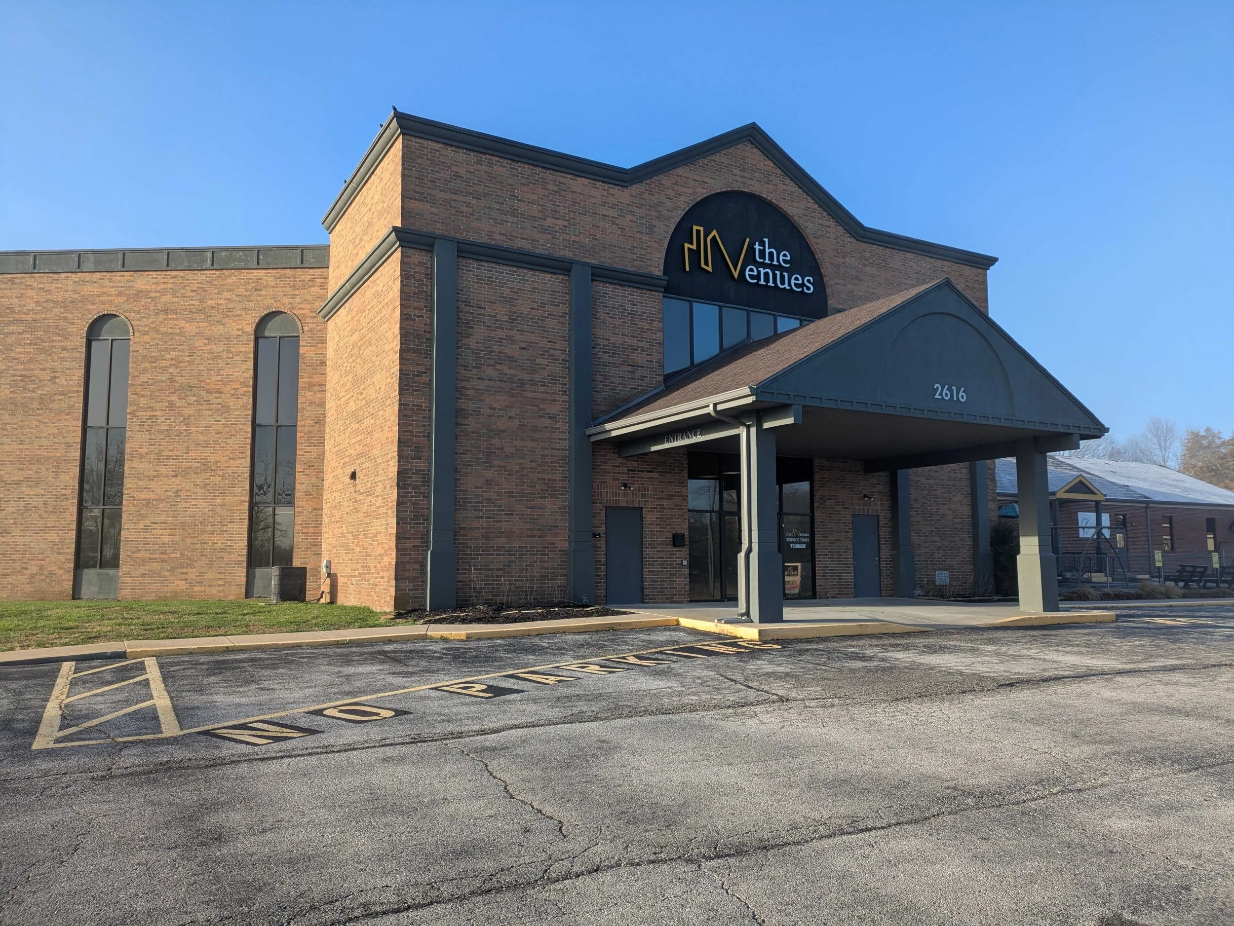 A brick building with a sign that reads 'the venues' and the address 2616. There is a covered entrance with a small roof and parking spaces in the foreground with yellow marking lines.