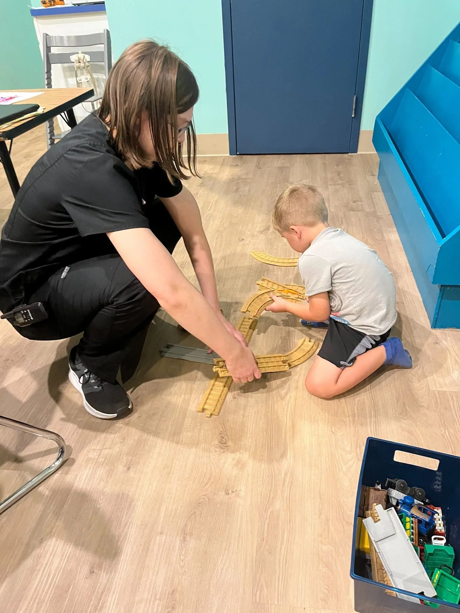A woman and a young boy kneeling on the floor playing with a wooden train set in a room with light wood flooring, a blue storage unit, and a table with chairs.