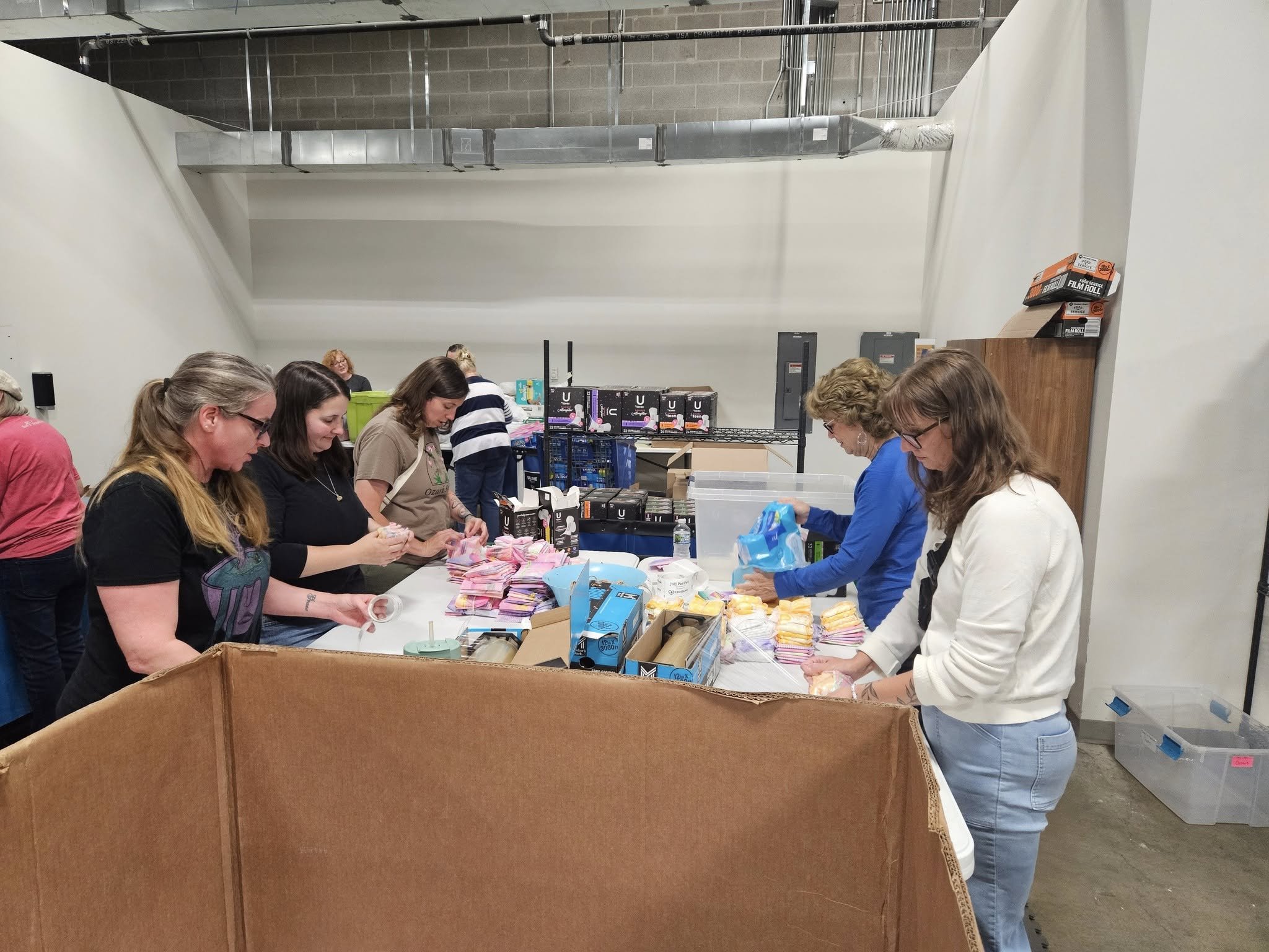 A group of people standing around a table sorting through various items, with boxes of supplies and grocery items on the table, likely involved in a volunteering or donation activity in an industrial space with exposed pipes and concrete walls.