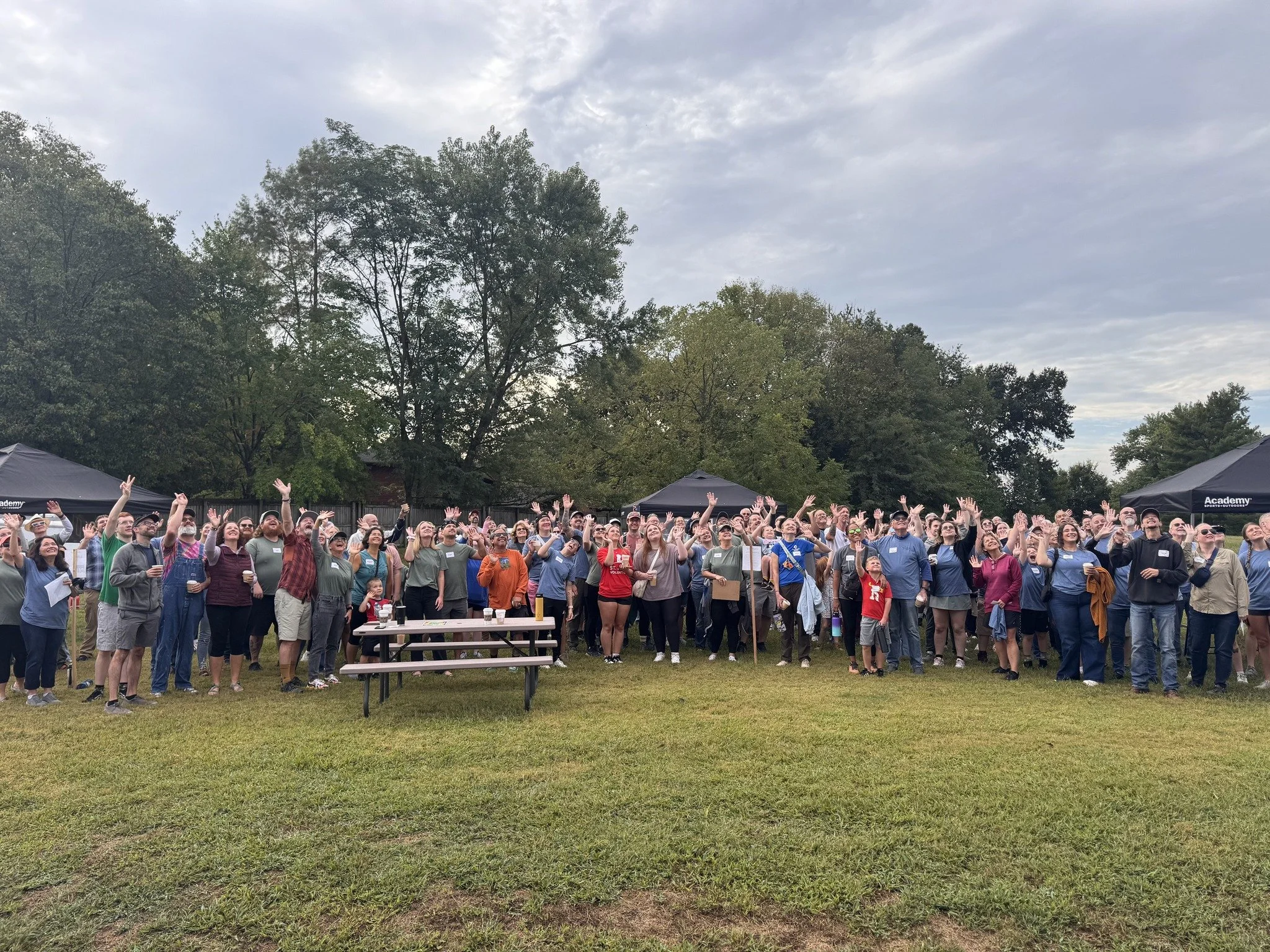 Large outdoor gathering of diverse people standing on grass, raising their hands in front of black tents with trees and cloudy sky in the background.