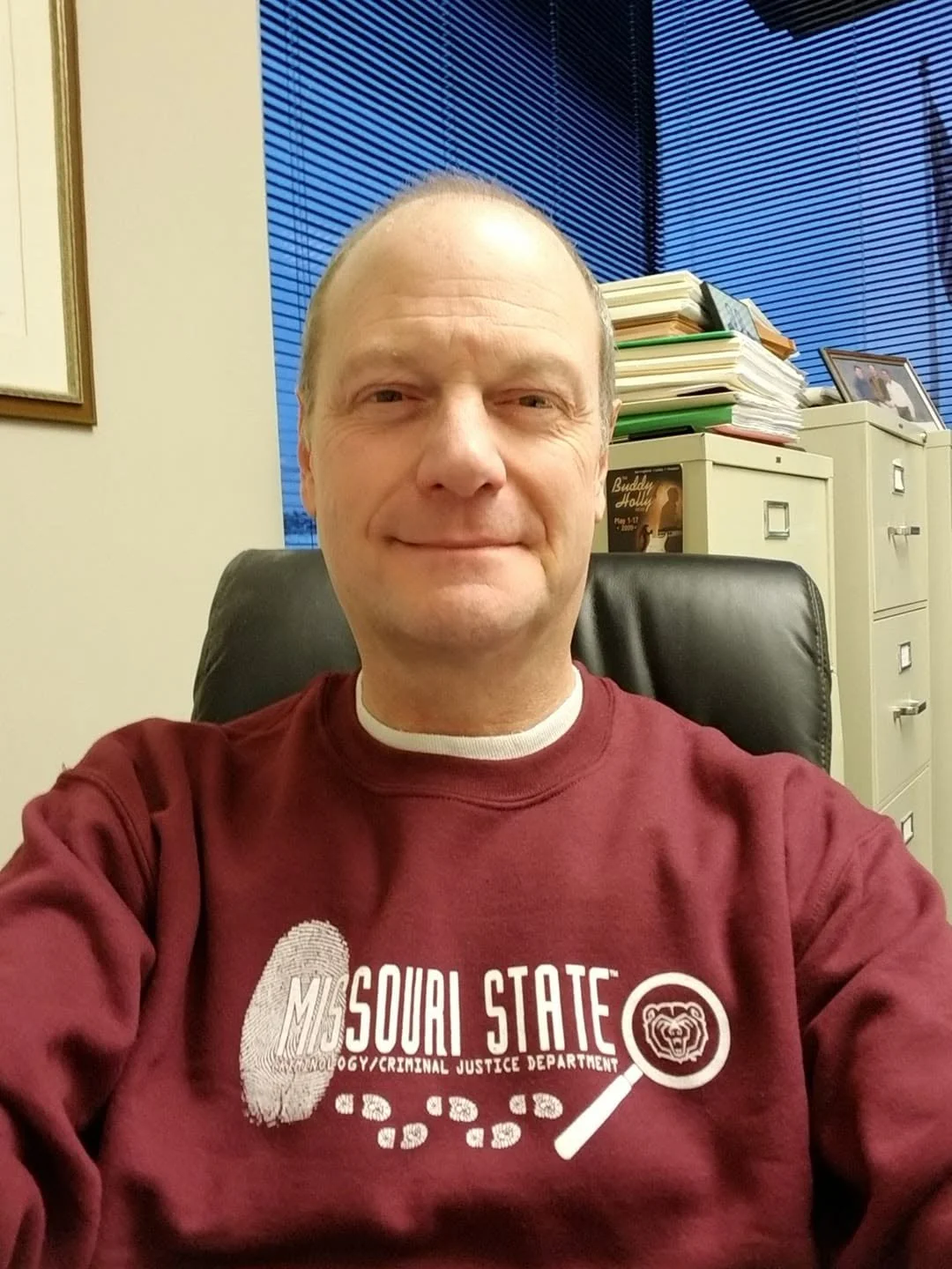 A man taking a selfie in an office, wearing a maroon Missouri State sweatshirt, with stacks of papers and a file cabinet in the background.
