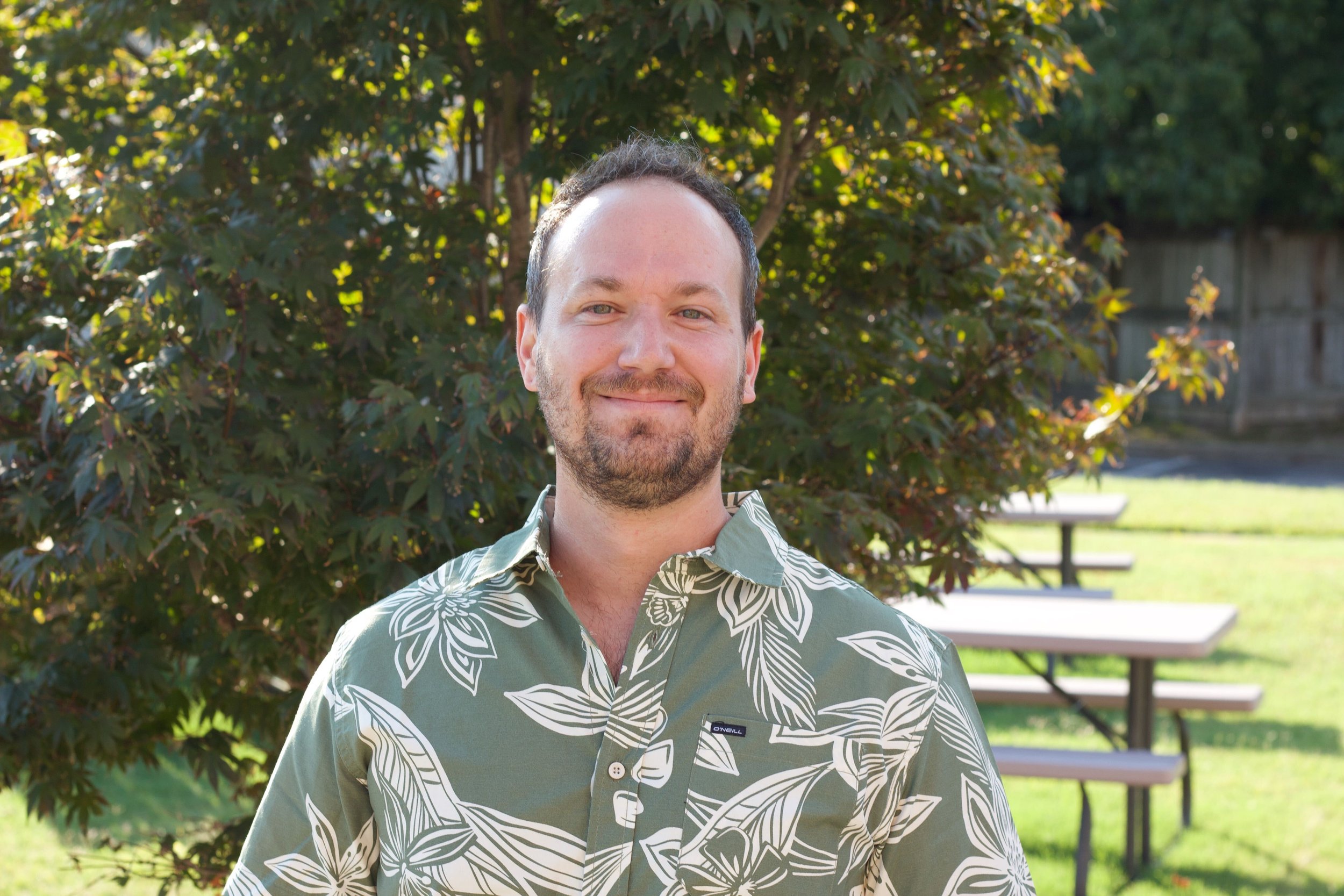 A man with short dark hair, a beard, and light skin is smiling and standing outdoors in front of a green leafy bush with sunlight filtering through.
